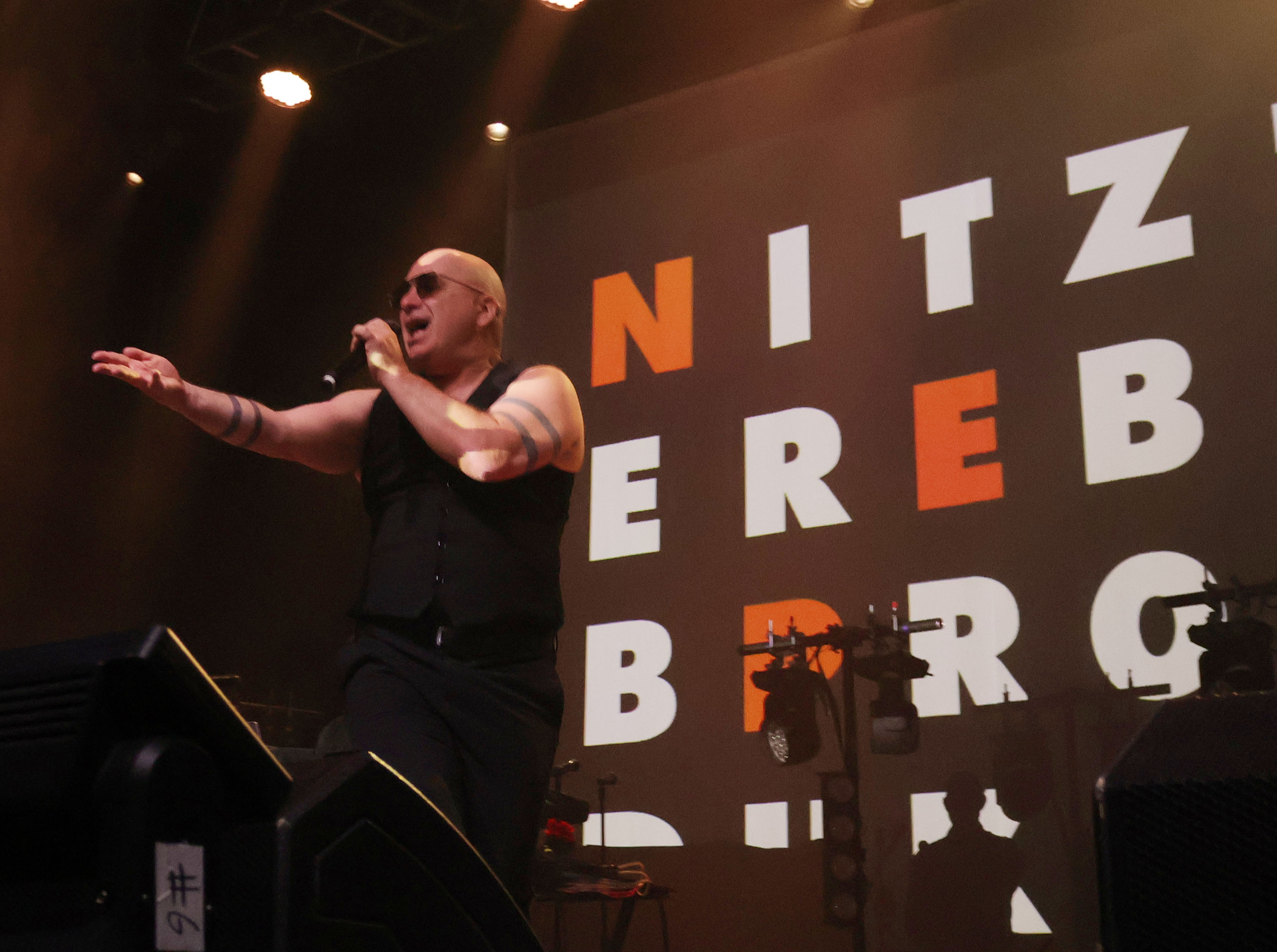 A singer in a black outfit performing energetically on stage with the backdrop displaying the name 'Nitzer Ebb' in bold letters.