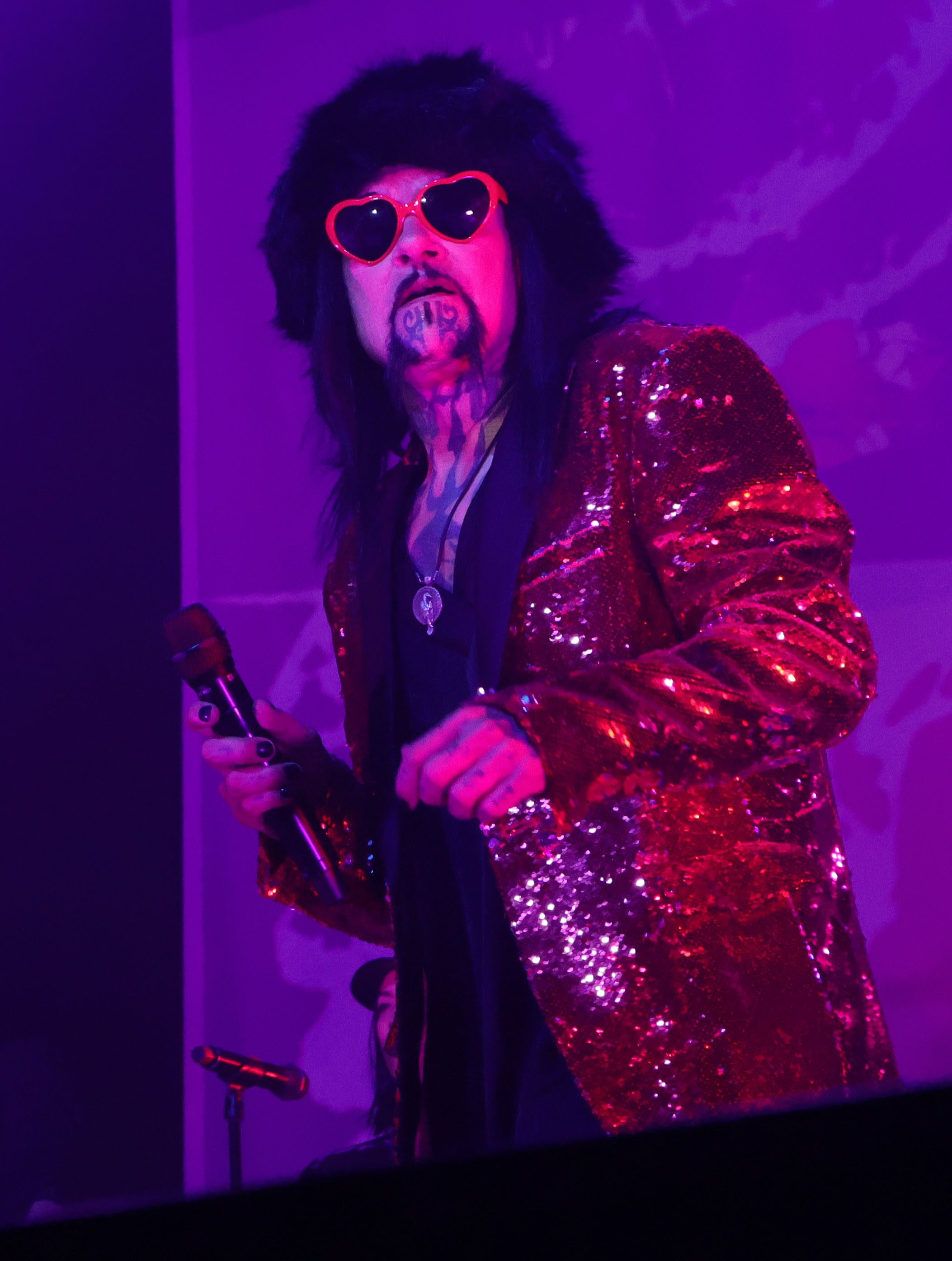 A performer wearing heart-shaped sunglasses and a glittery red jacket stands on stage, with purple lighting in the background.
