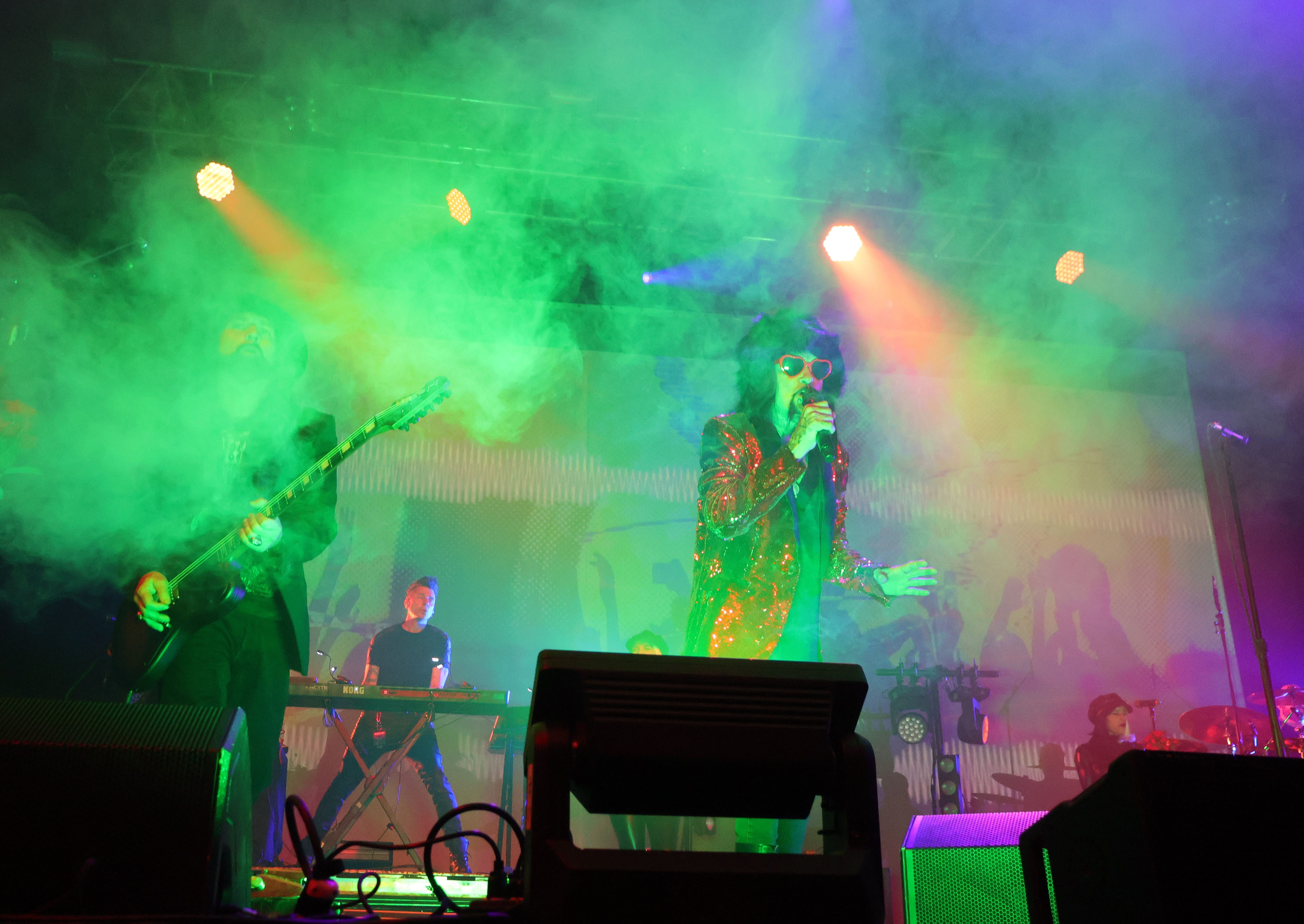 A live performance scene featuring musicians on stage with vibrant green and purple lighting and smoke effects. The lead singer, wearing sunglasses and a shiny jacket, is engaging with the audience, while the guitarist and keyboardist are in the background.