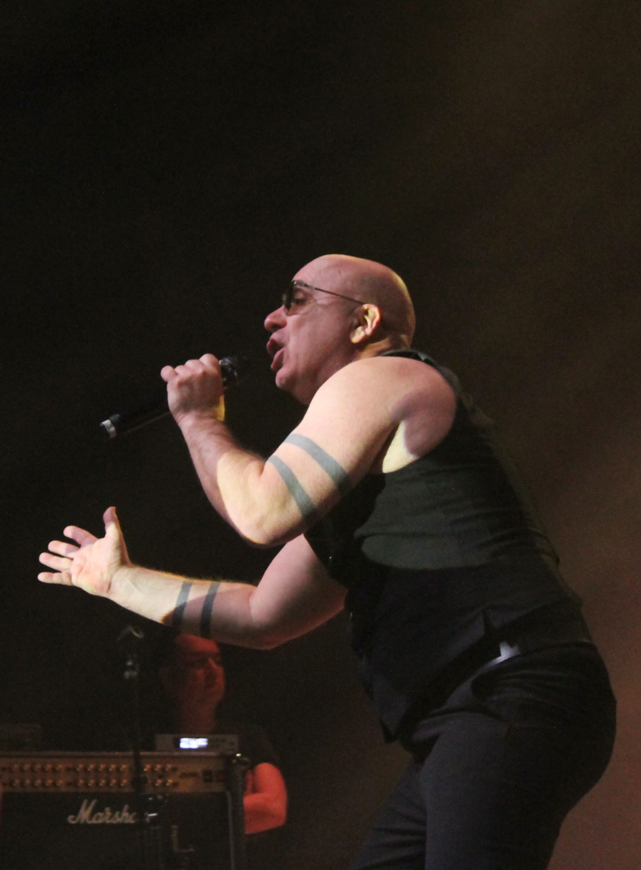 A performer passionately singing into a microphone during a concert, with dramatic stage lighting in the background.