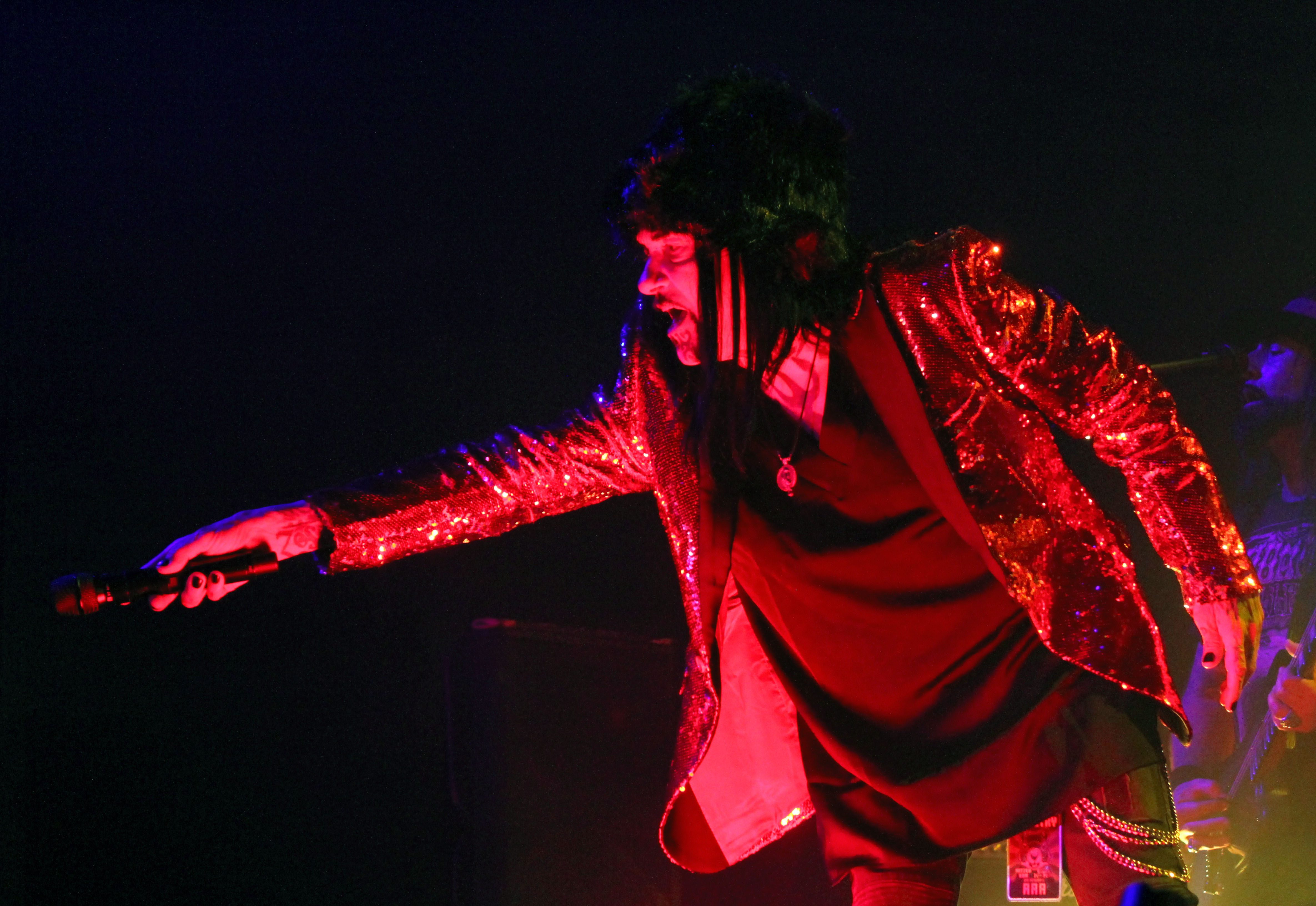 A performer on stage wearing a shiny red sequined jacket, reaching out with a microphone in hand, during a music concert.
