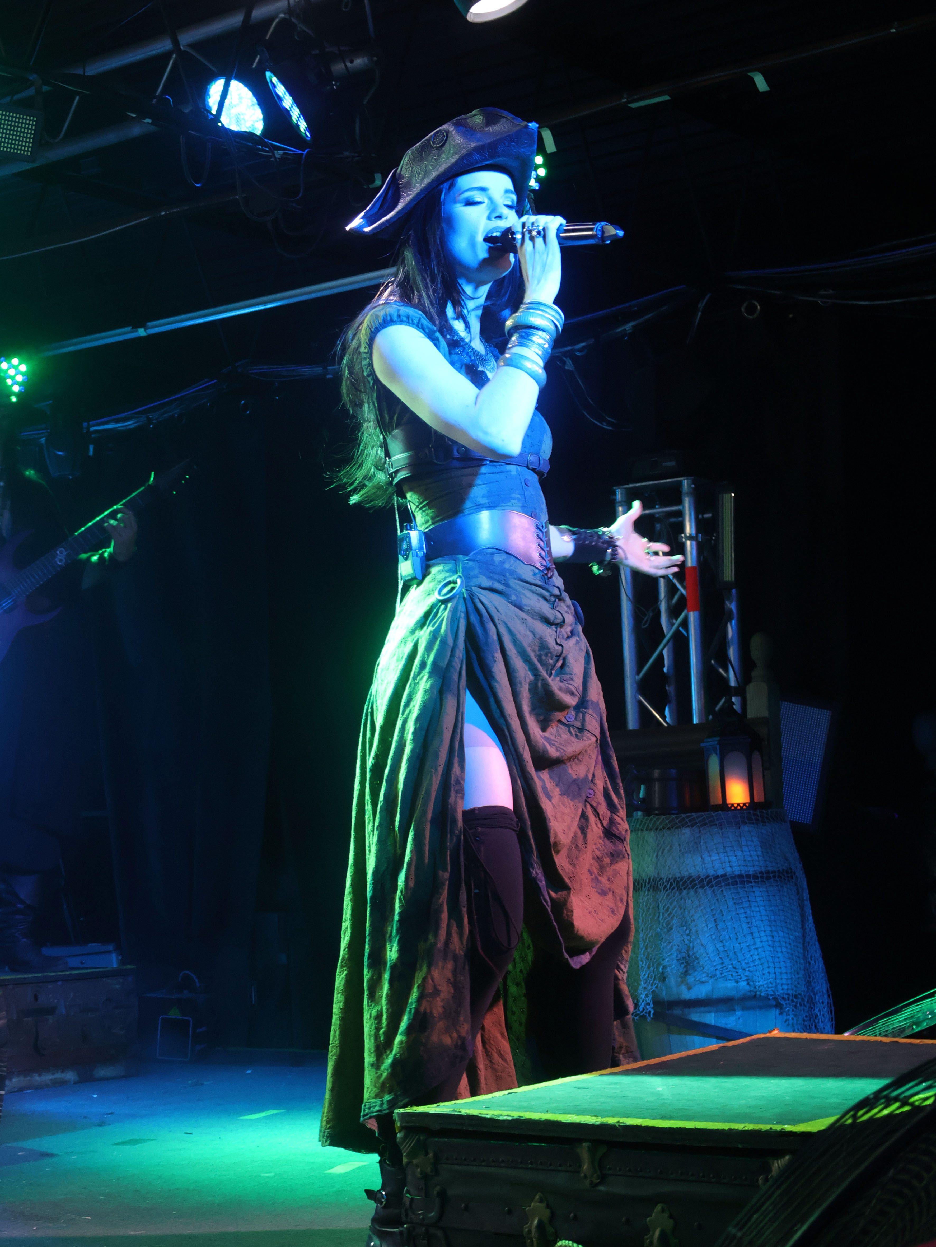 A singer in a pirate-themed outfit passionately performing on stage, holding a microphone with a dramatic pose, while colorful stage lights illuminate the scene.