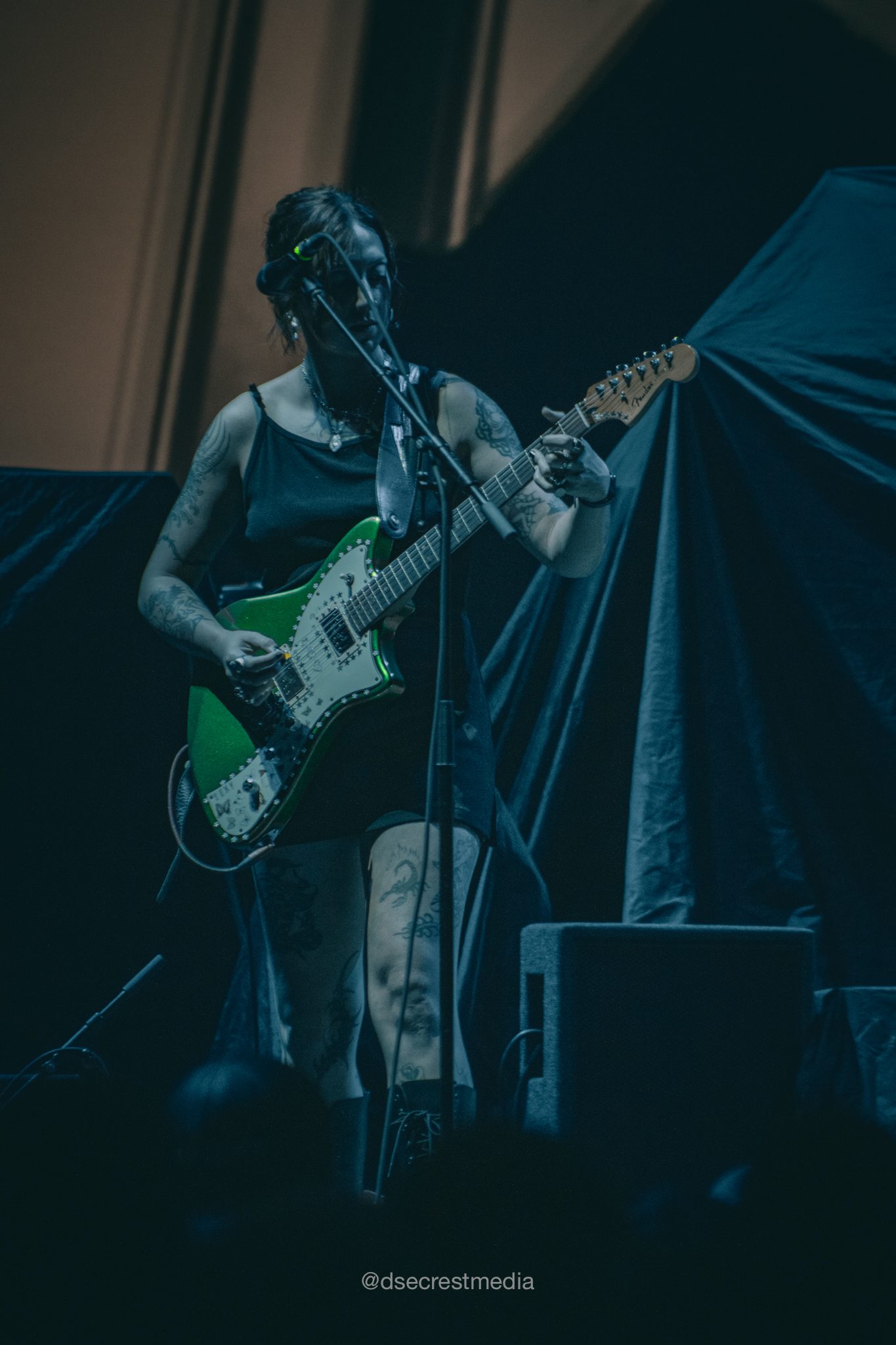 A musician performing on stage, playing a green electric guitar while wearing a black outfit. The focus is on her upper body, with visible tattoos on her arms.