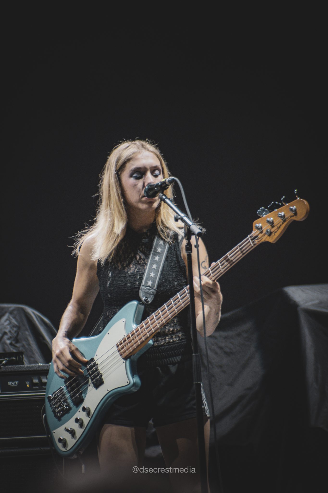 A female bassist performs on stage, singing into a microphone while playing a turquoise bass guitar during a live music event.