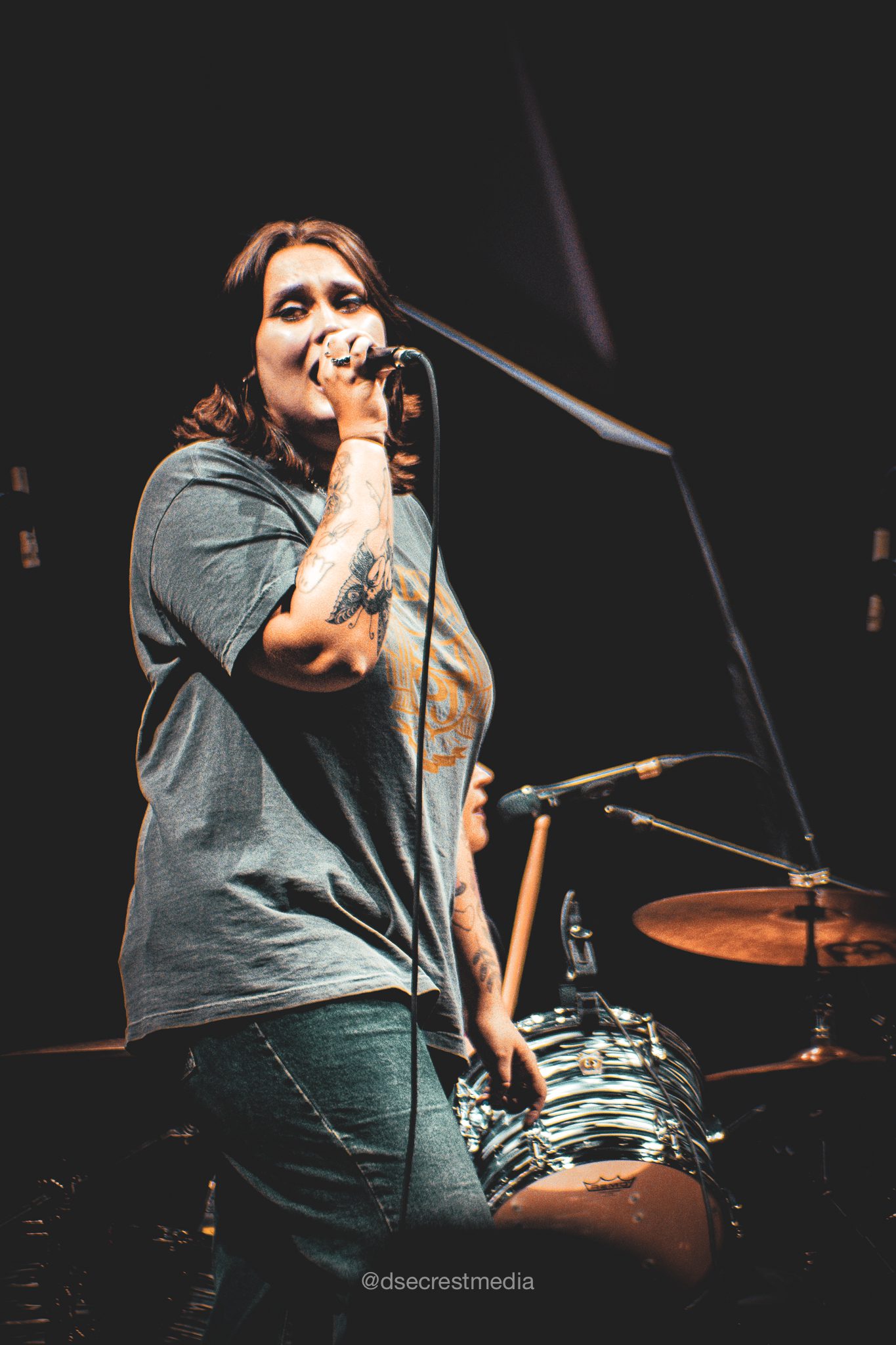 A female vocalist passionately singing into a microphone on stage, wearing a casual gray t-shirt, with band equipment visible in the background.
