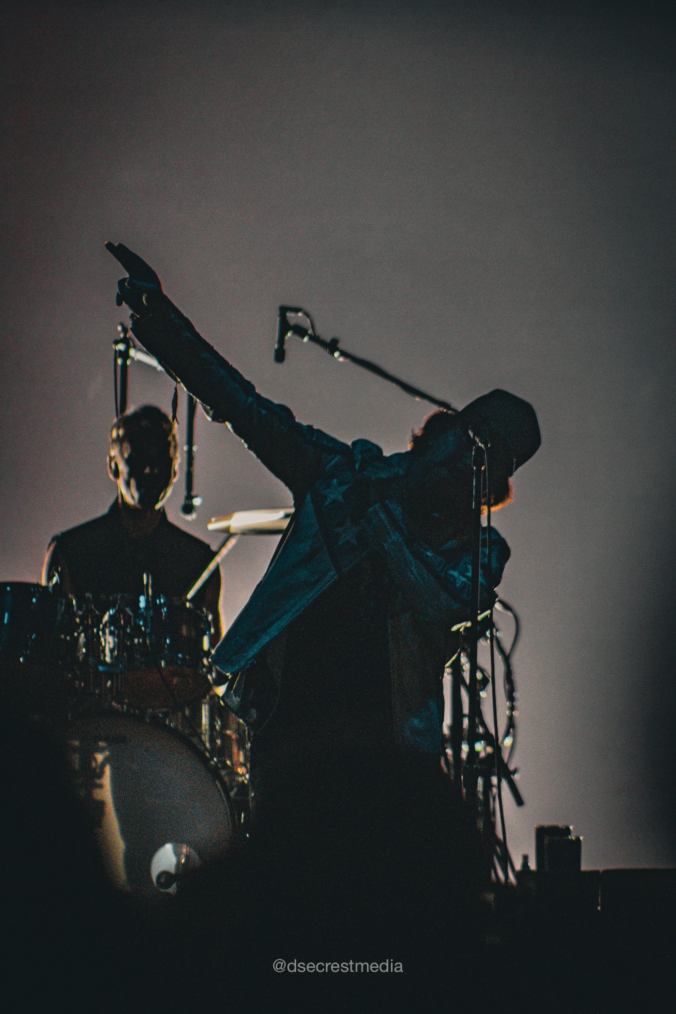 Silhouette of a musician on stage with a raised arm, accompanied by a drummer in the background, during a live concert performance.
