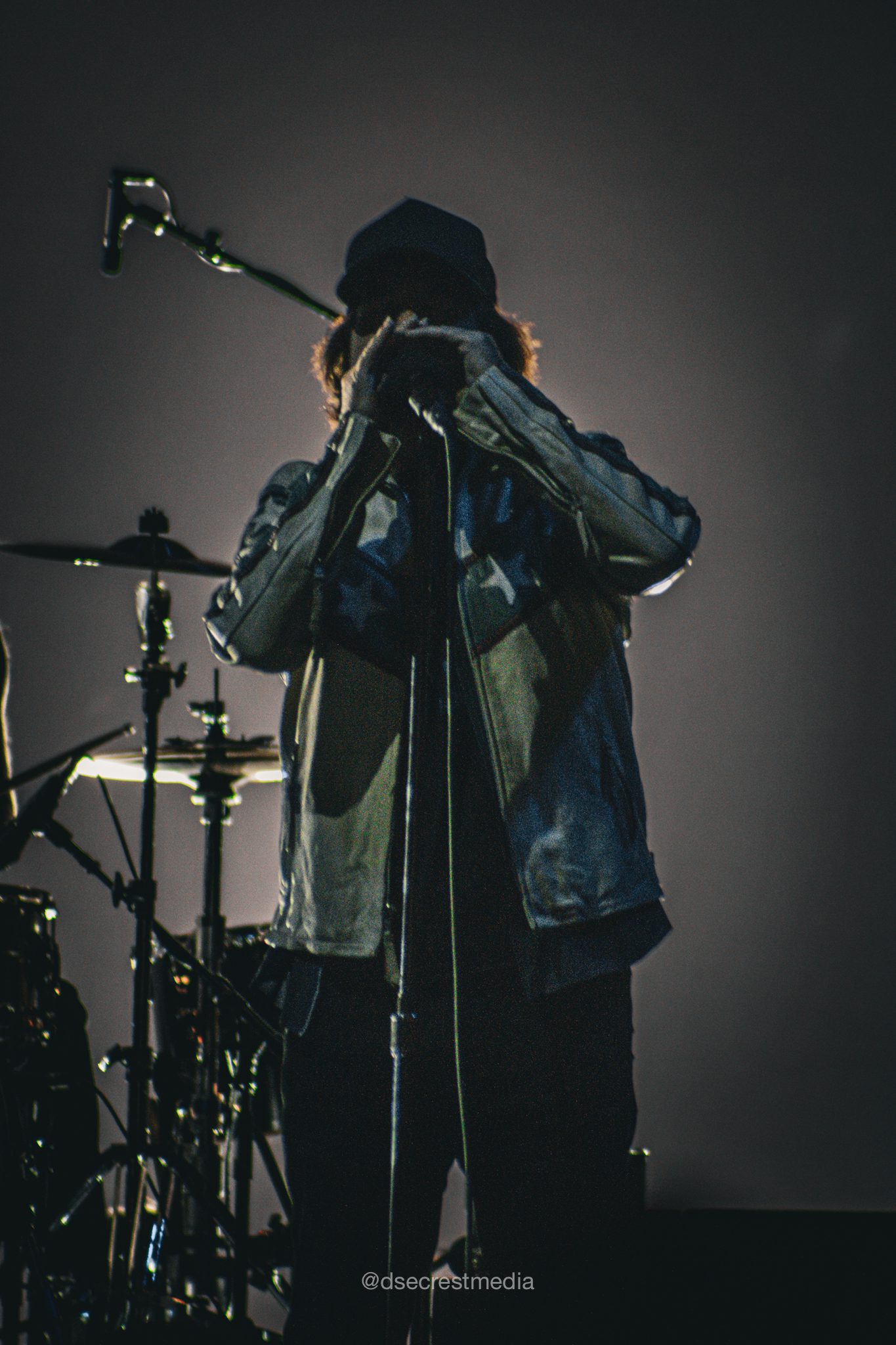 A silhouette of a musician singing into a microphone with a drum set in the background, illuminated by stage lights.