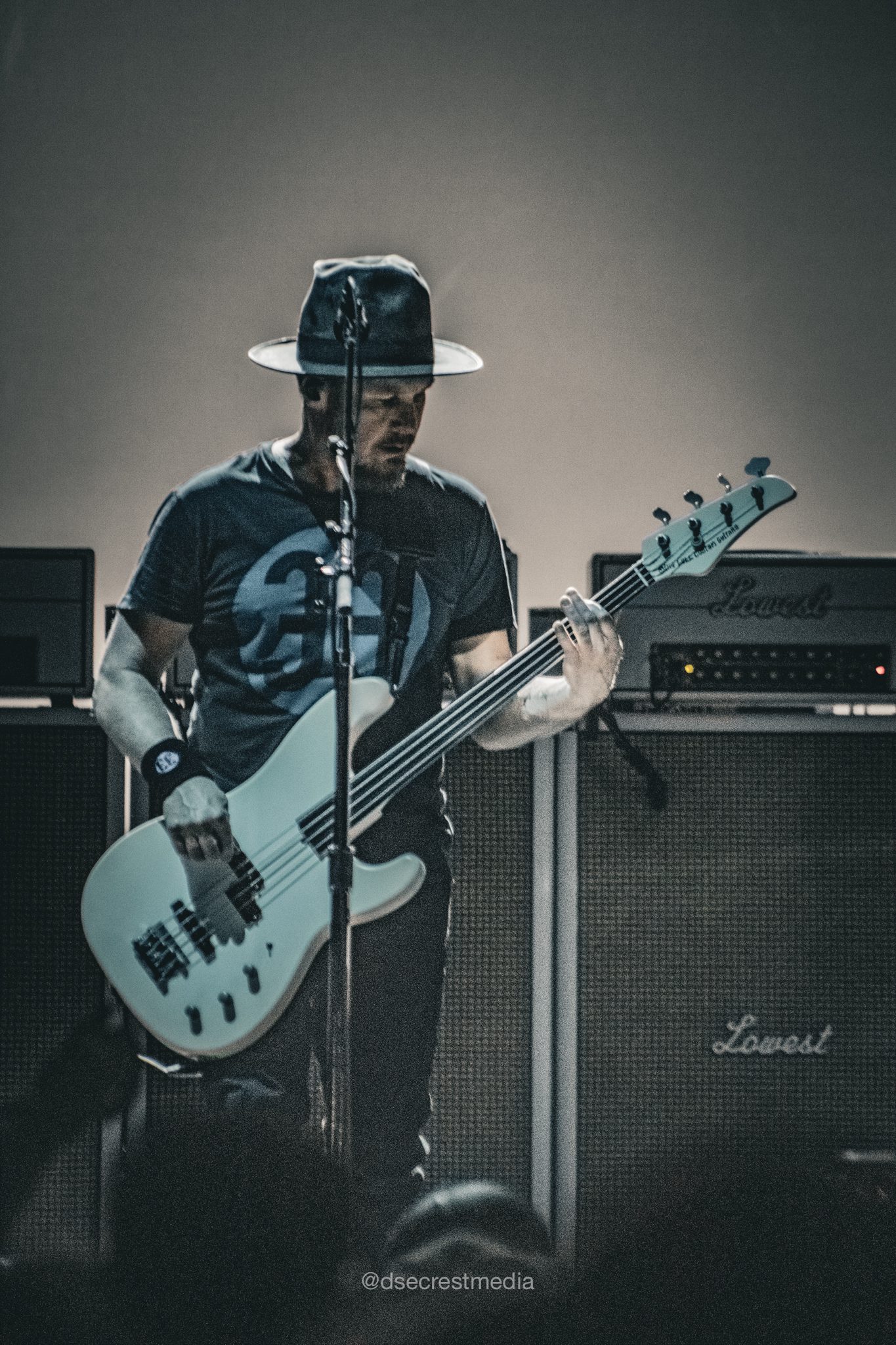 A musician playing a white bass guitar on stage during a concert, wearing a hat and a graphic t-shirt, with amplifier stacks in the background.