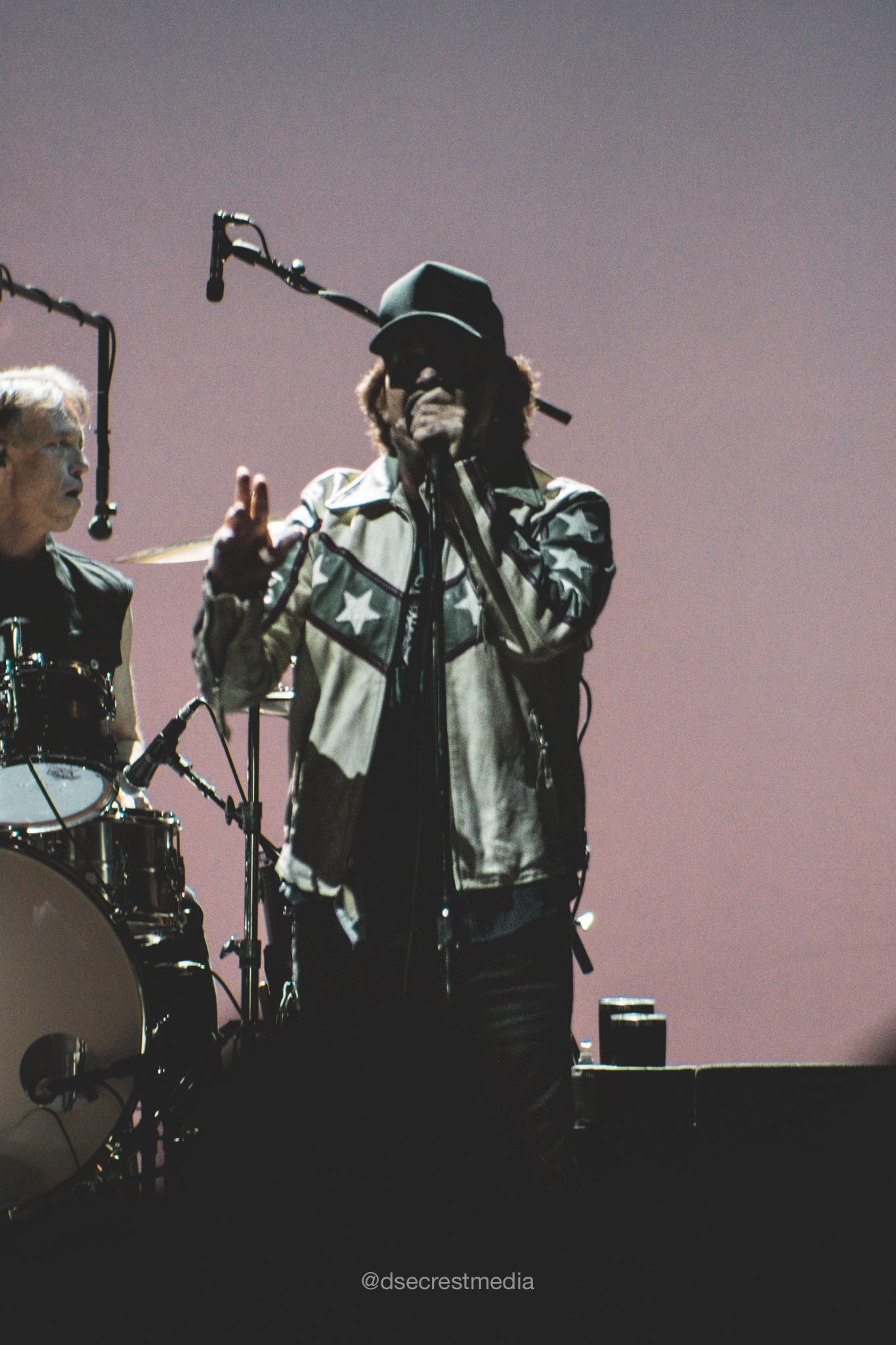 A lead singer in a star-patterned jacket performing on stage with a drummer in the background, in a vibrant lighting setup.