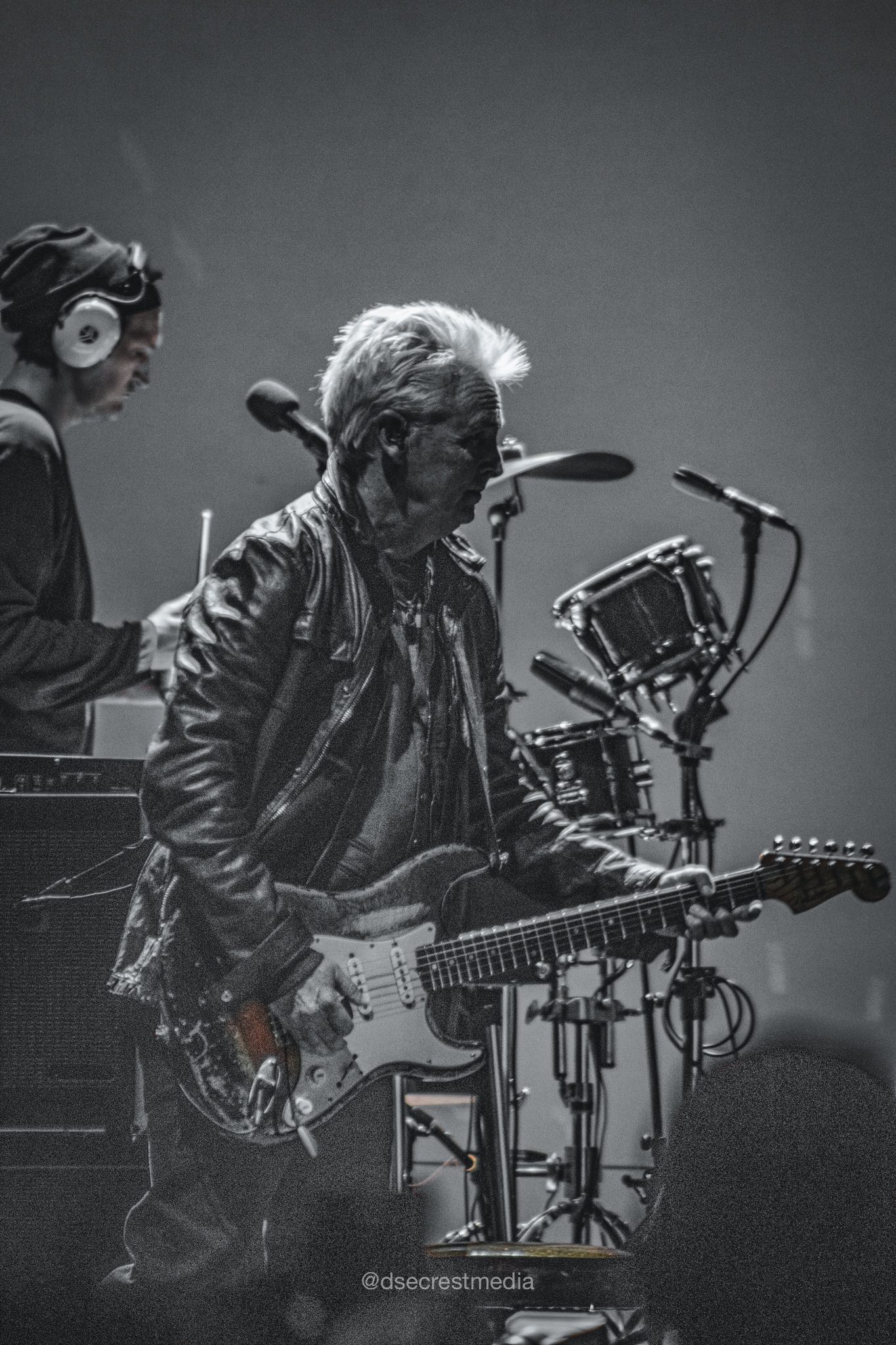 A guitarist in a leather jacket plays an electric guitar on stage, with a drummer visible in the background. The image is in black and white, capturing the energy of a live music performance.
