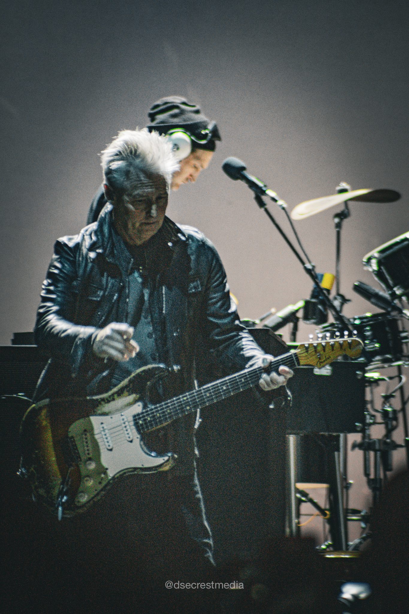 A guitarist performing on stage, wearing a black leather jacket, with a serious expression, as a drummer is visible in the background.