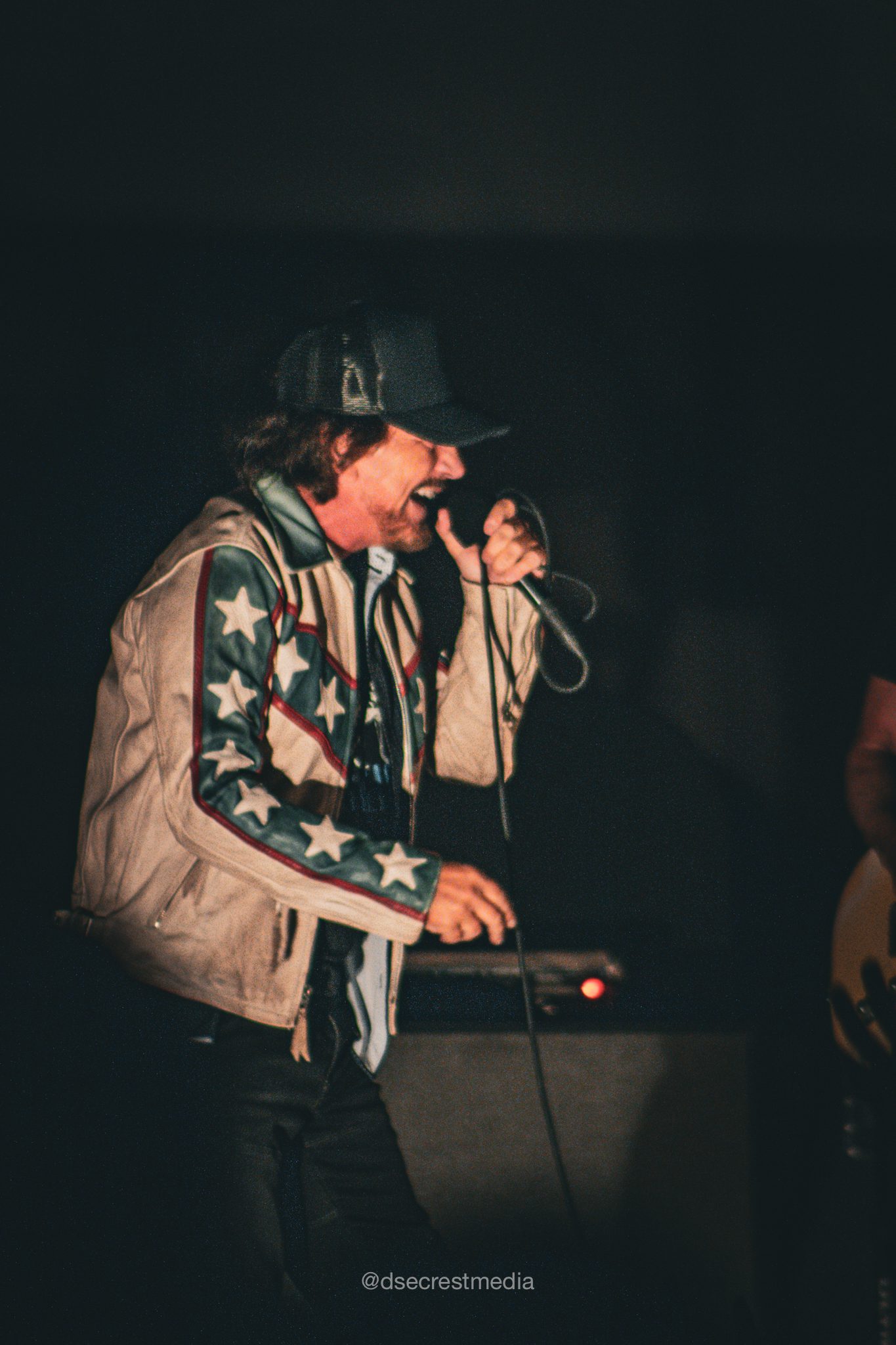 A male singer passionately performing on stage, wearing a star-patterned jacket and holding a microphone, with a blurred crowd in the background.