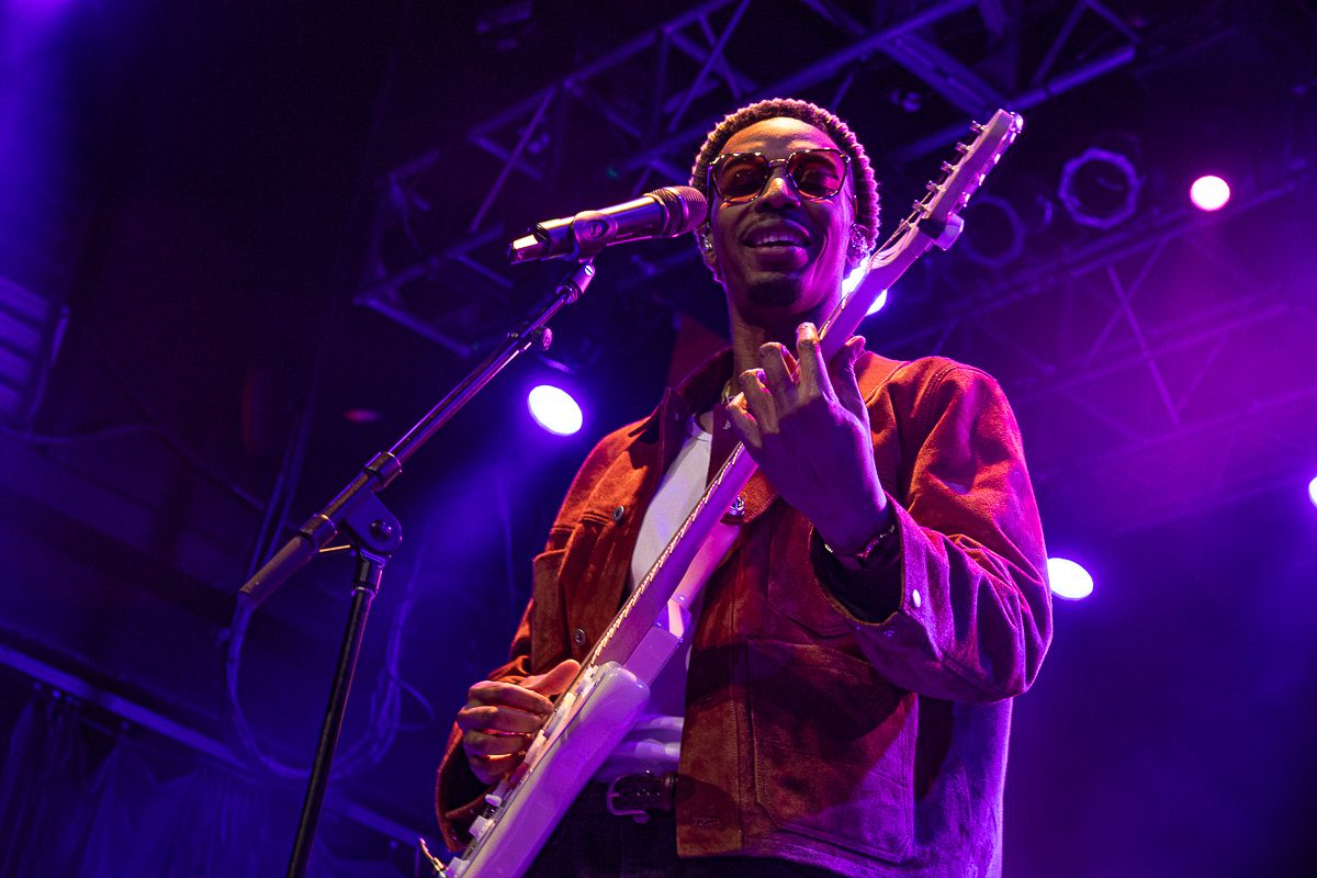 Anees performing on stage with a guitar, wearing sunglasses and a brown jacket, with colorful stage lighting in the background.