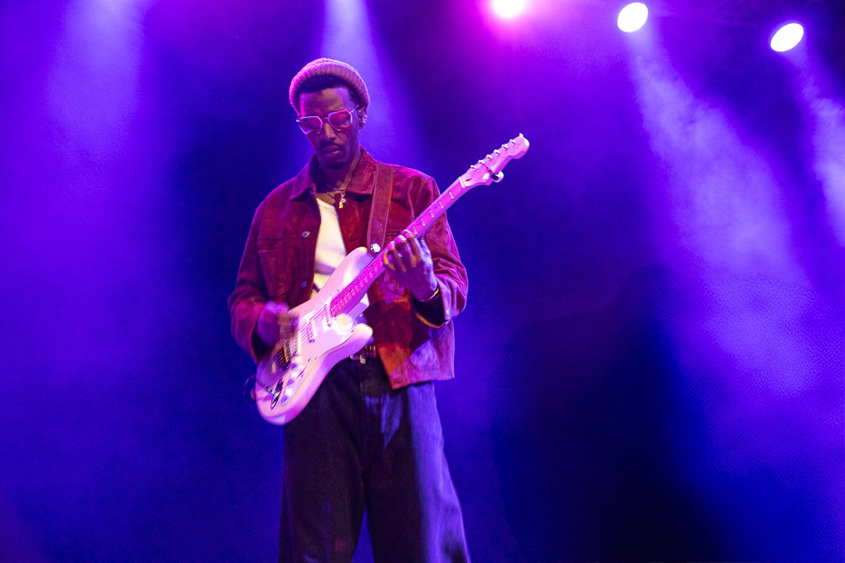 A musician performing on stage with a guitar, dressed in a red jacket and a beanie, under purple stage lights.