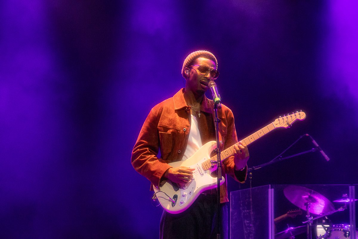 A musician performing on stage with a guitar, wearing sunglasses and a brown jacket, against a purple-lit background.