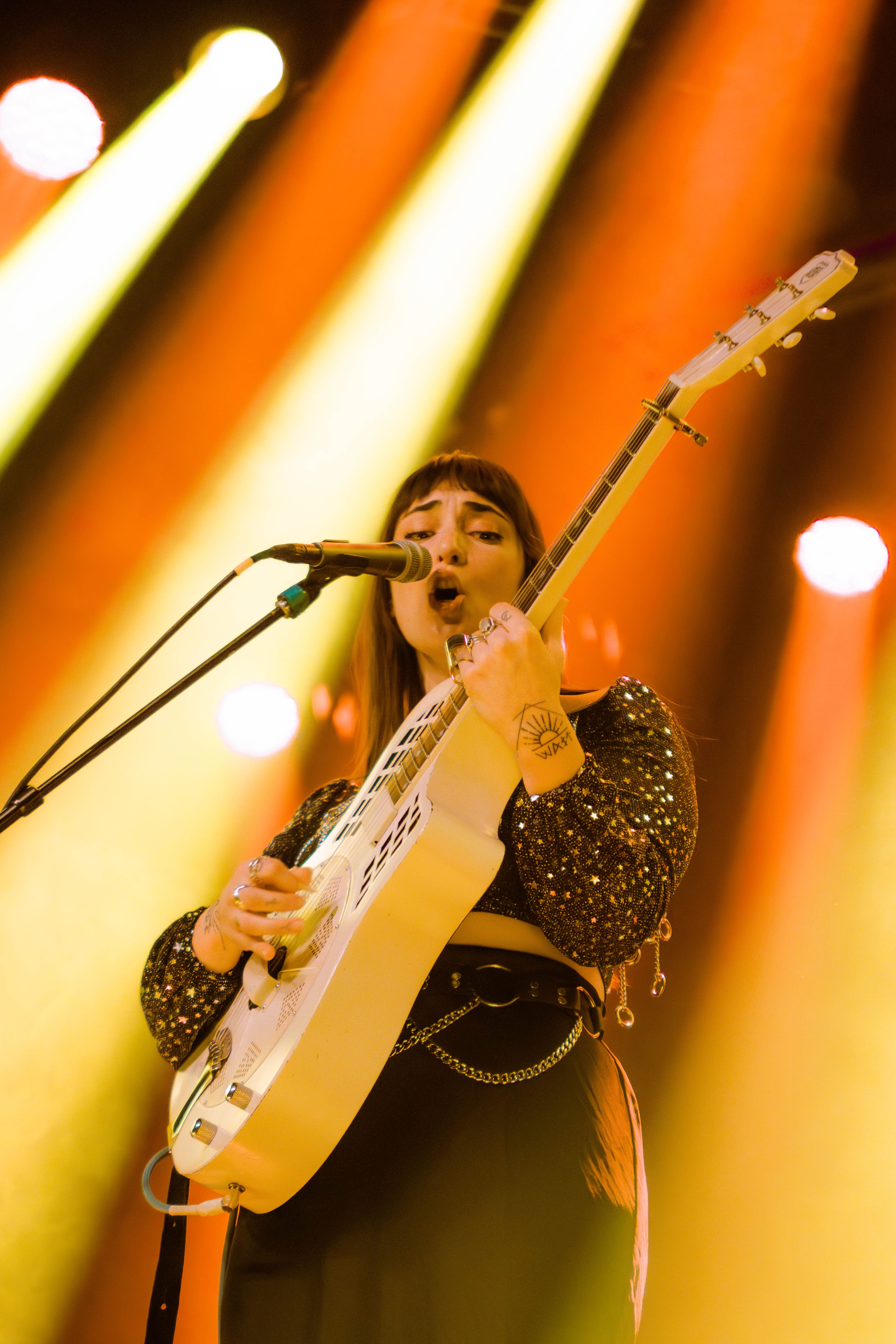 A singer-songwriter performing on stage with a resonator guitar, wearing a sparkly black top, under bright yellow stage lights.