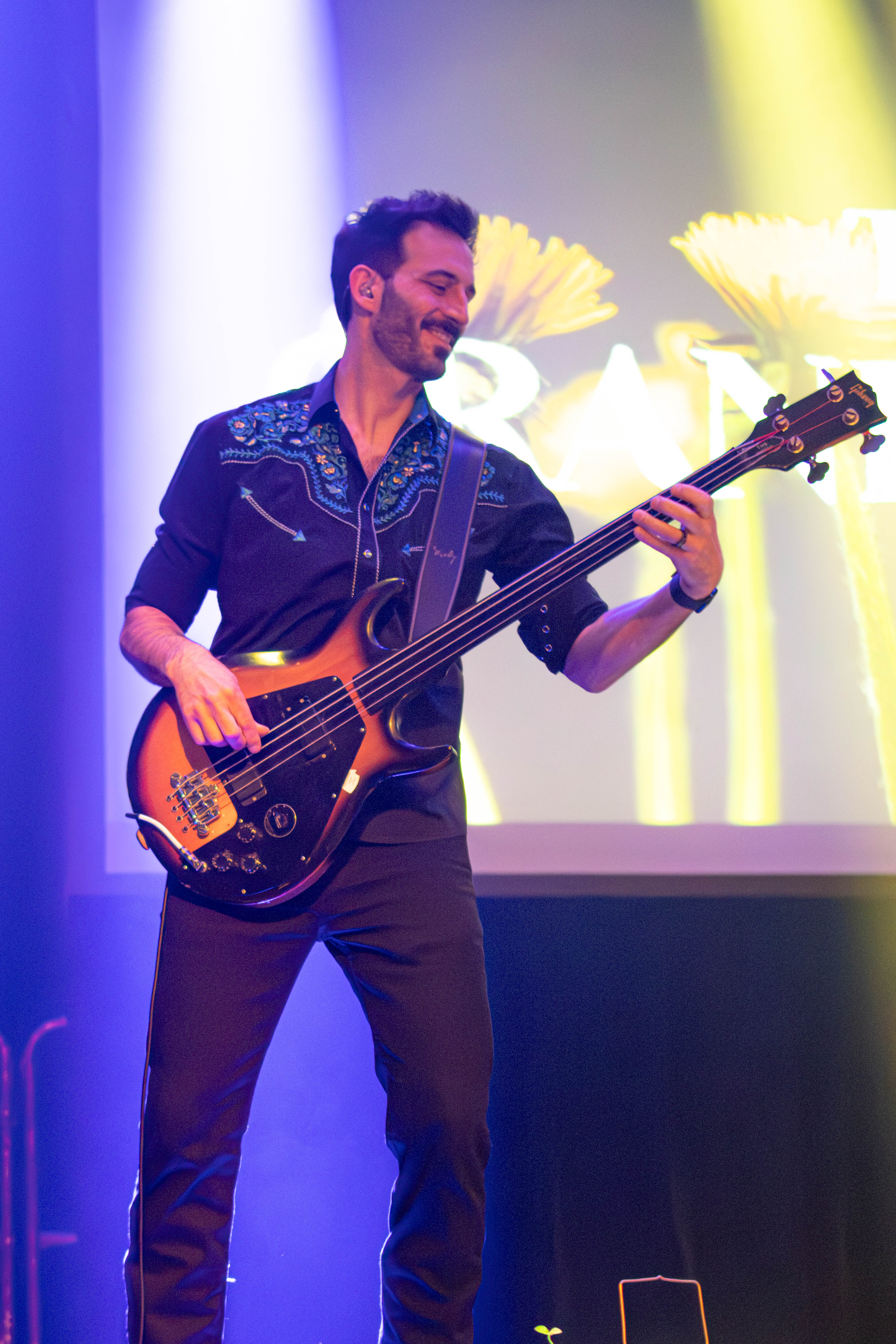 A bassist performing on stage, smiling while playing an electric bass guitar under colorful stage lights.
