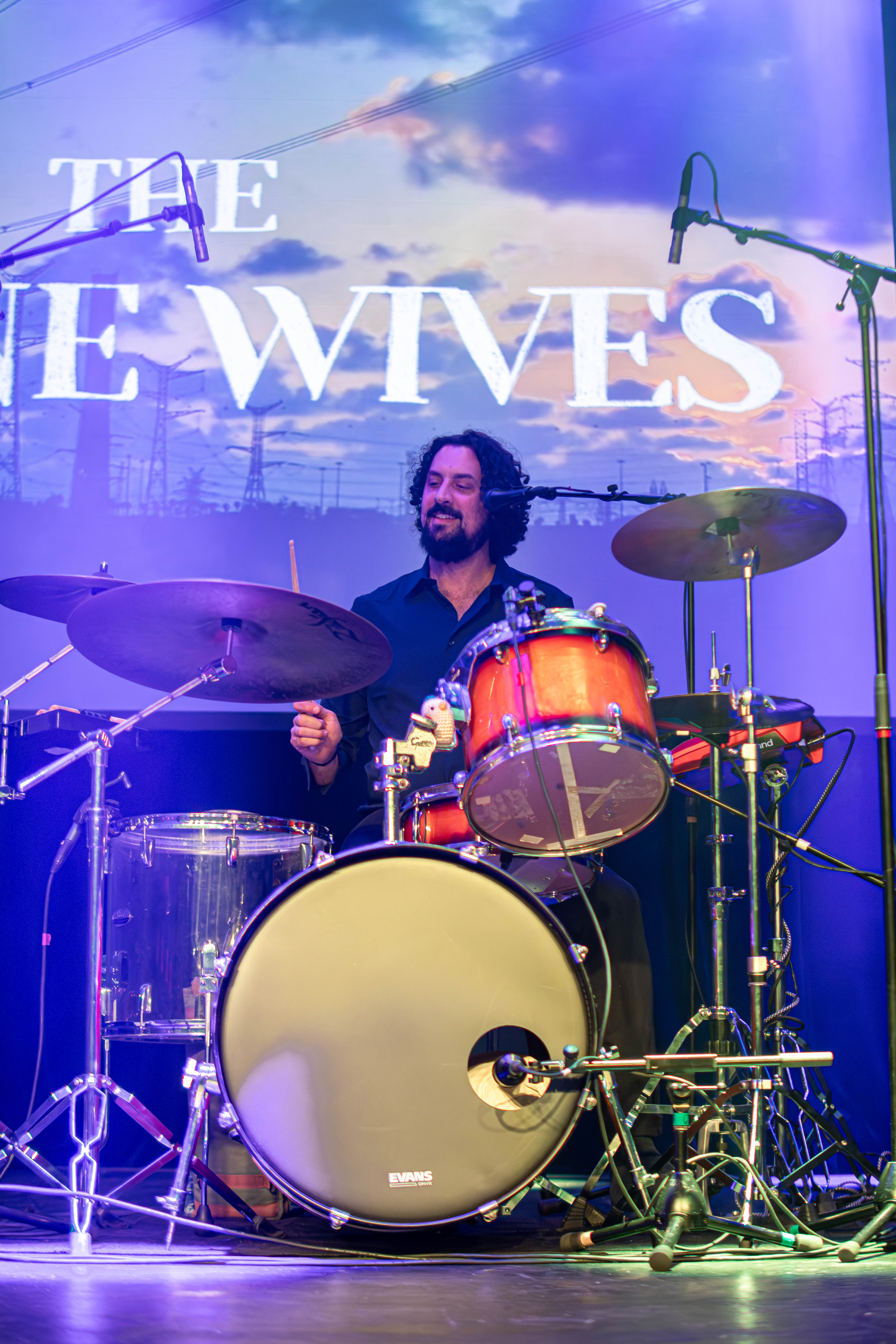 A drummer performing on stage, smiling while playing a drum set with bright stage lights in the background.