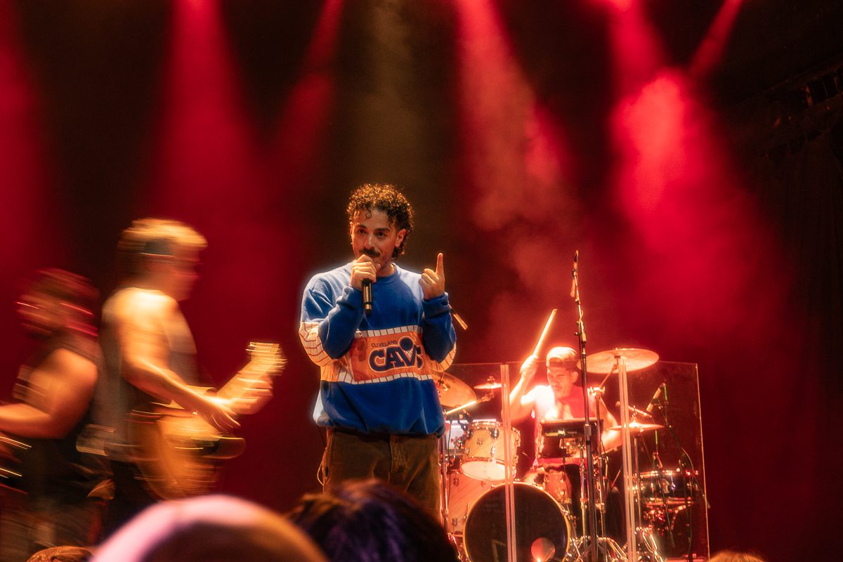 Anees performing on stage with a microphone, dressed in a blue sweatshirt, surrounded by band members and red stage lights.