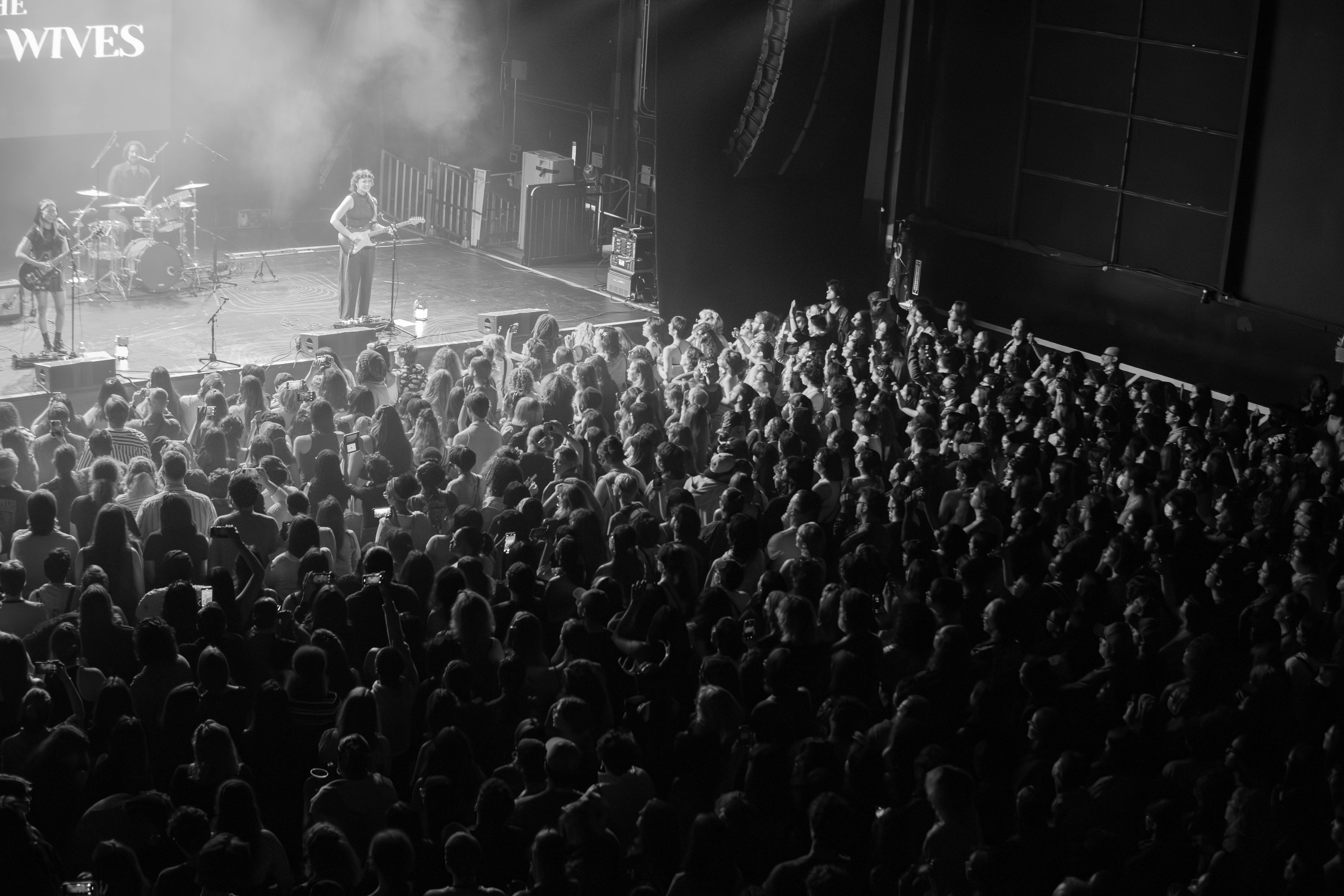 A live performance by The Crane Wives on stage, with the band members visible and a large audience in the foreground, captured in black and white.