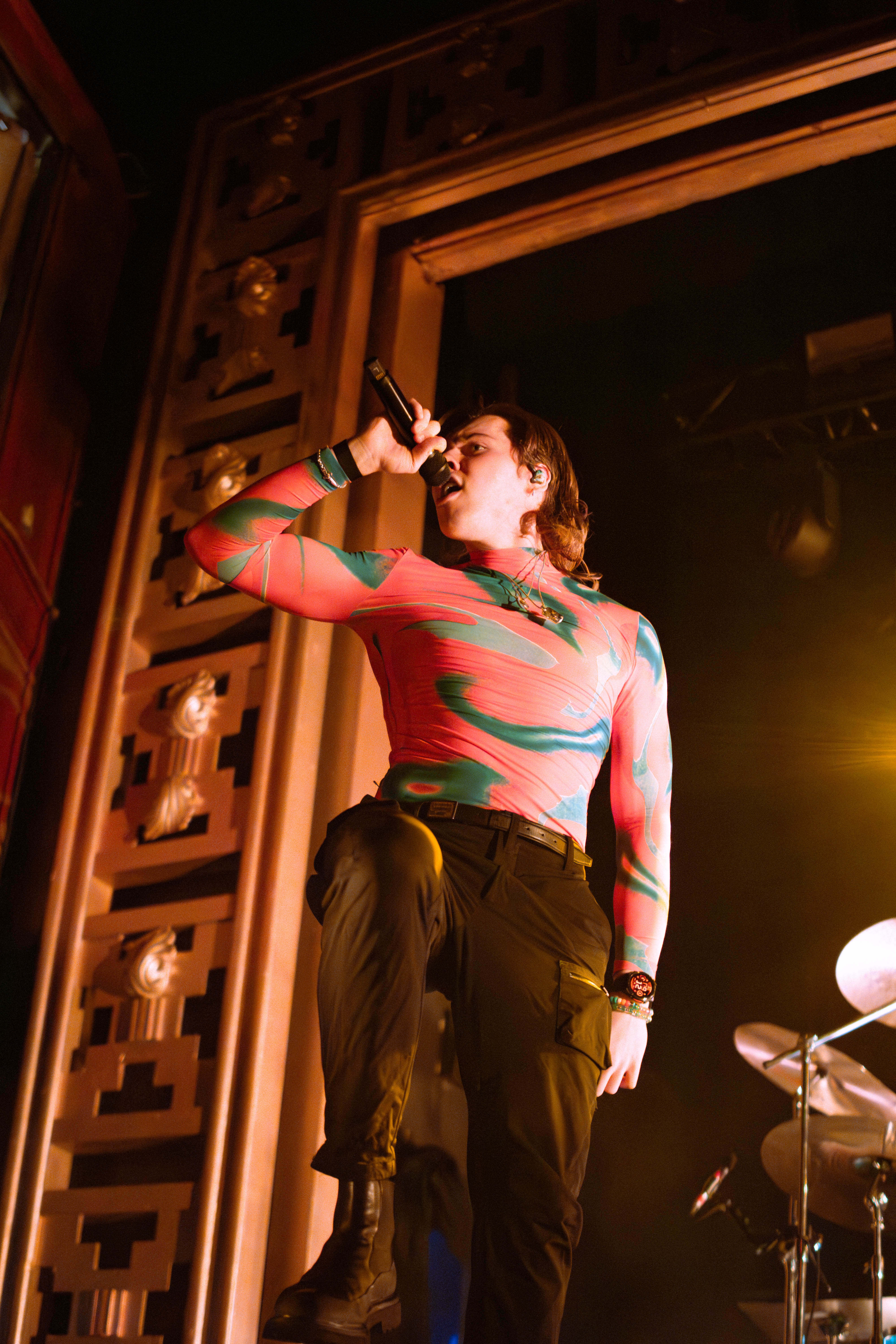 A performer singing into a microphone on stage, wearing a colorful long-sleeve shirt and black pants, with a musical setup visible in the background.