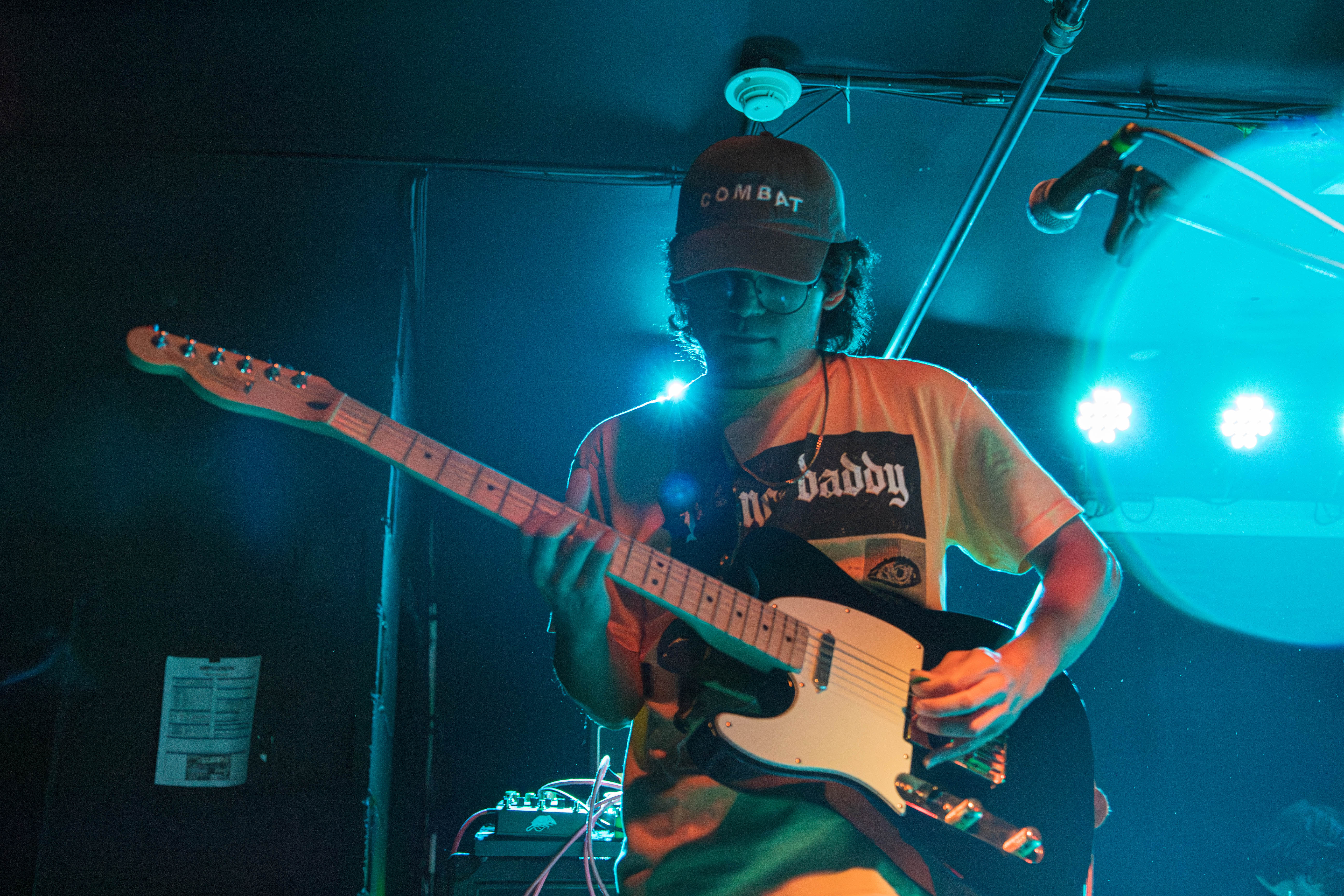 A musician playing an electric guitar under colorful stage lights, wearing a 'COMBAT' cap and a graphic t-shirt.