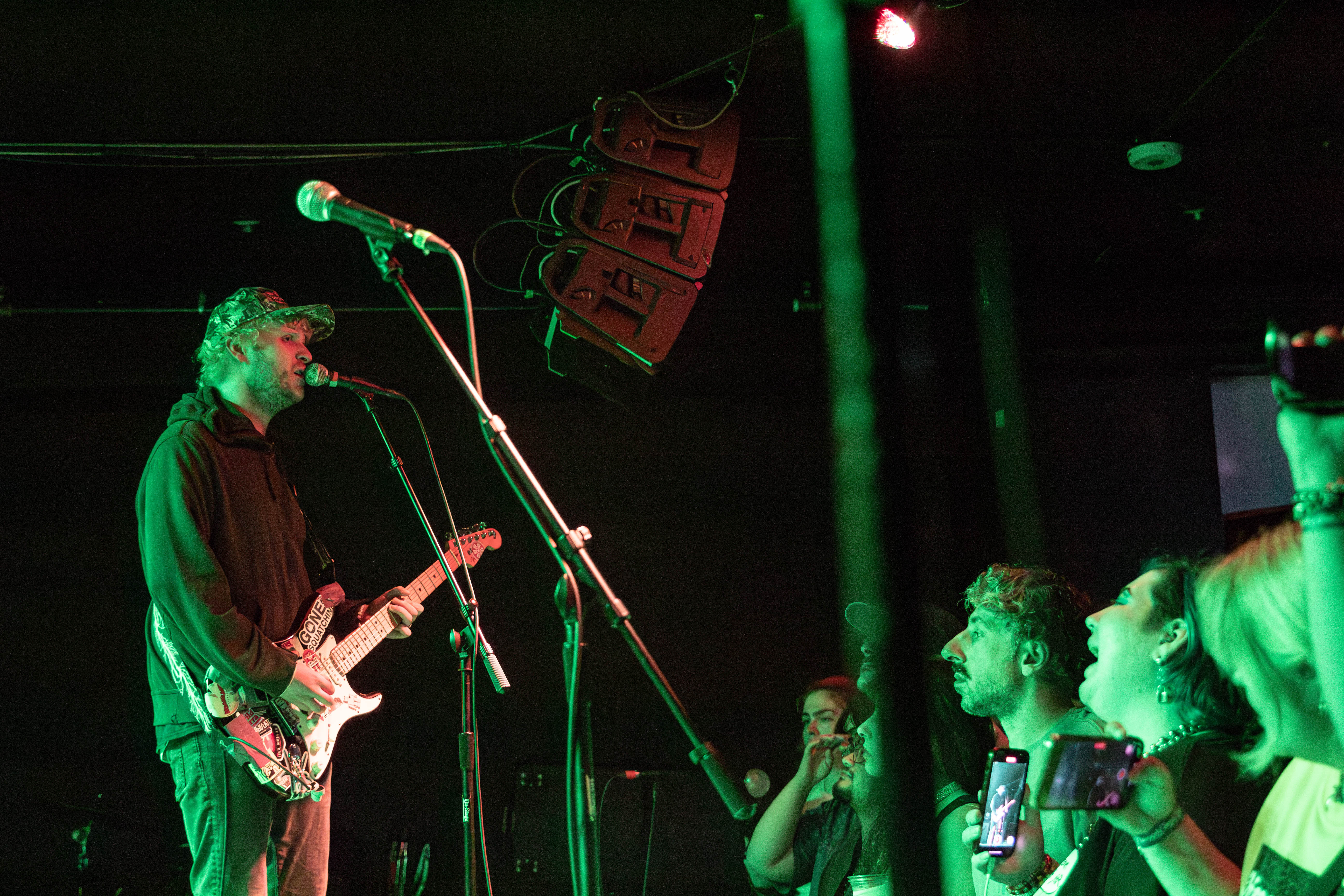A musician performing on stage with a microphone and electric guitar, while an engaged audience watches closely, illuminated by green lighting.