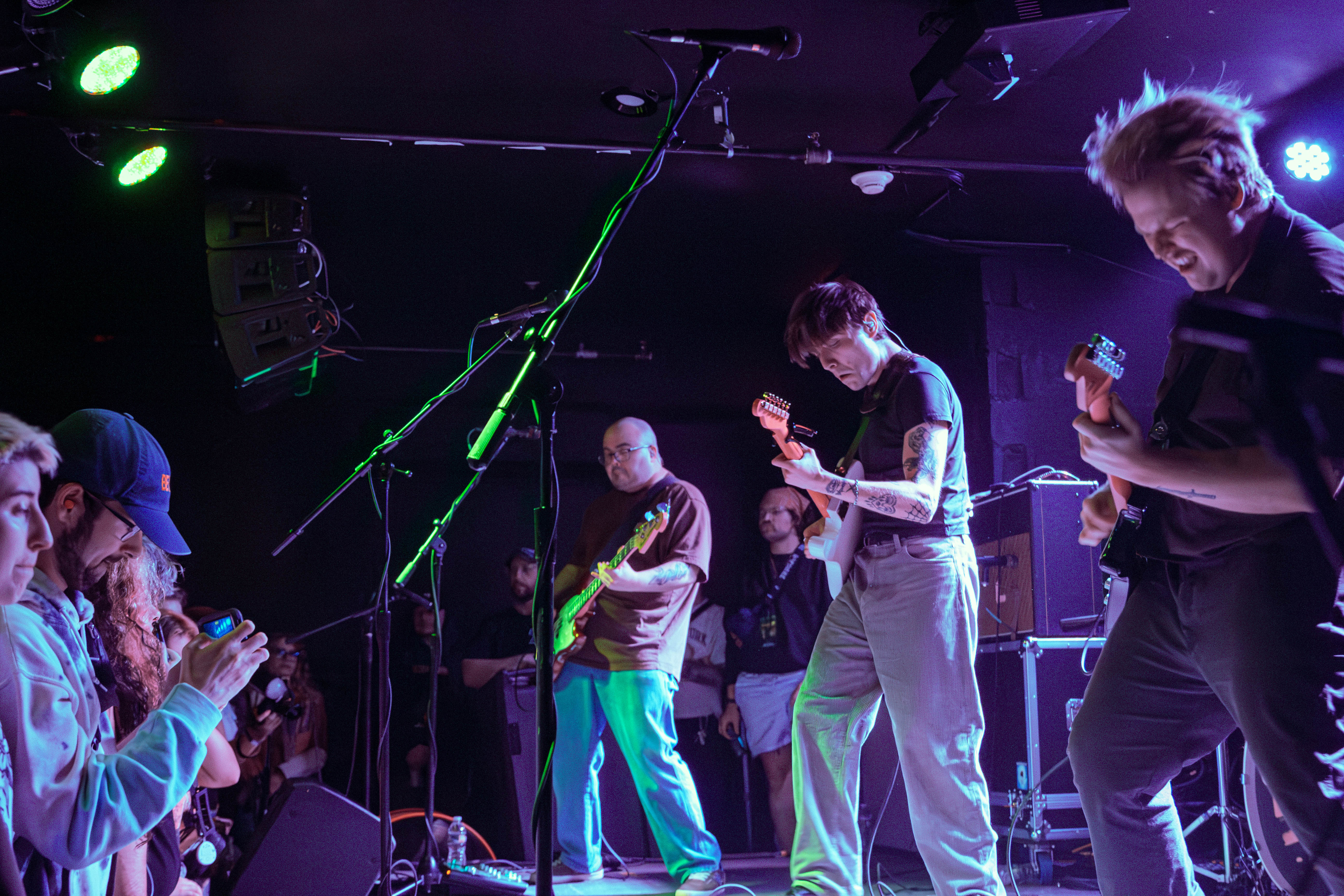 A live performance scene featuring a band on stage with band members playing instruments under colorful stage lights, while an engaged audience watches in the foreground.