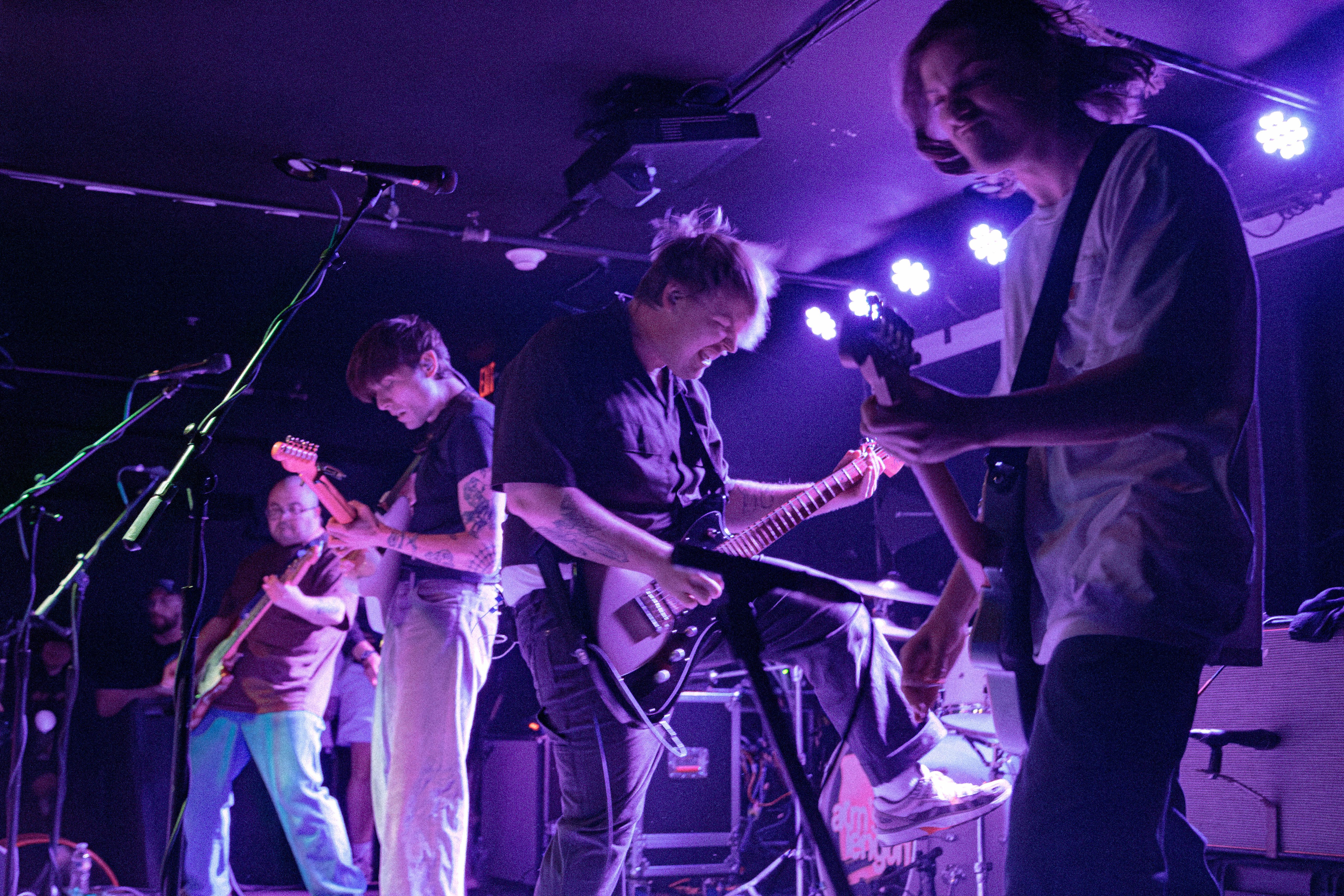 A live performance by the band Arm's Length, featuring four members on stage playing guitars and drums under purple lighting.