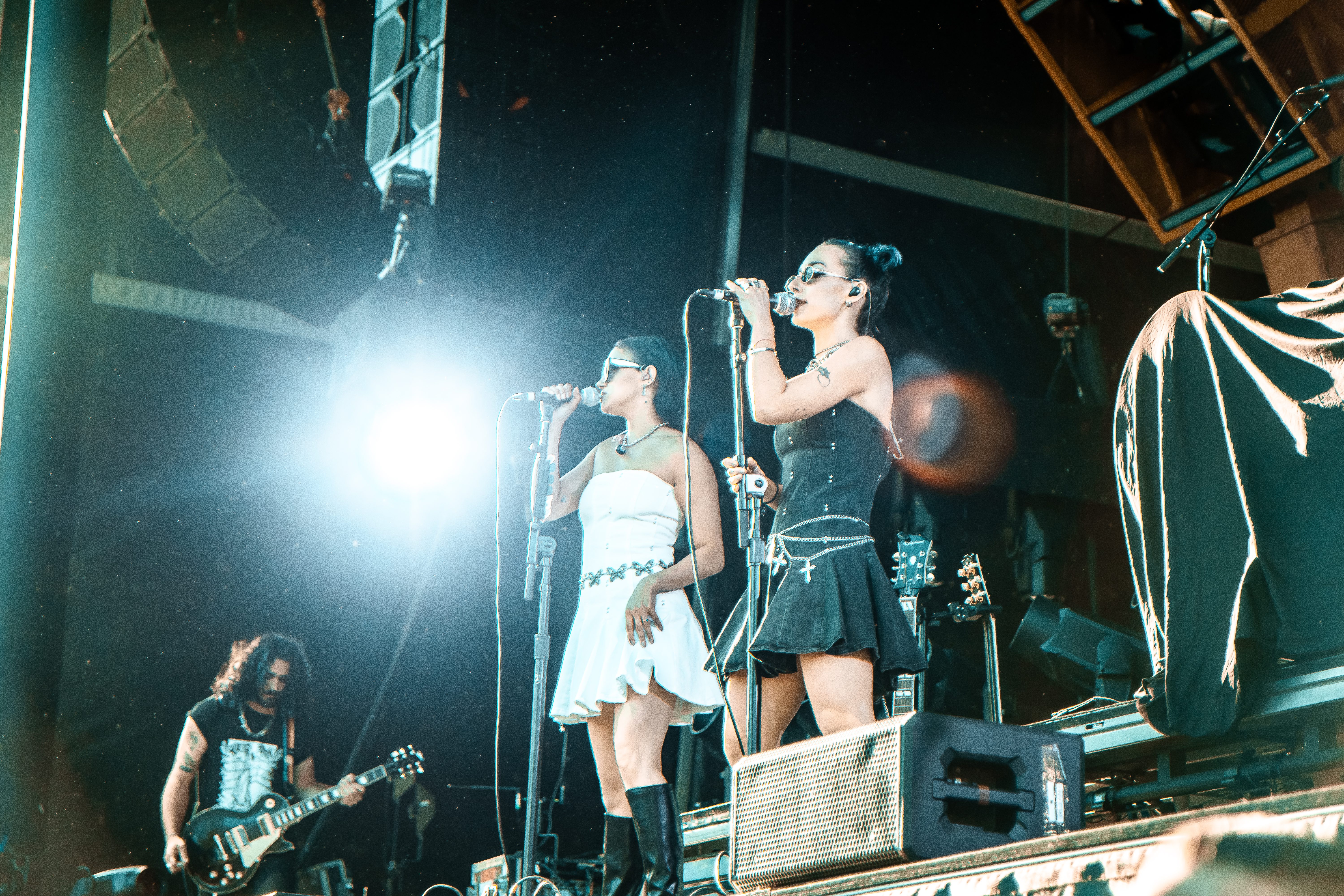 Two female singers performing on stage, one in a black dress and the other in a white dress, with microphones in hand and sunglasses on. A male guitarist is visible in the background.