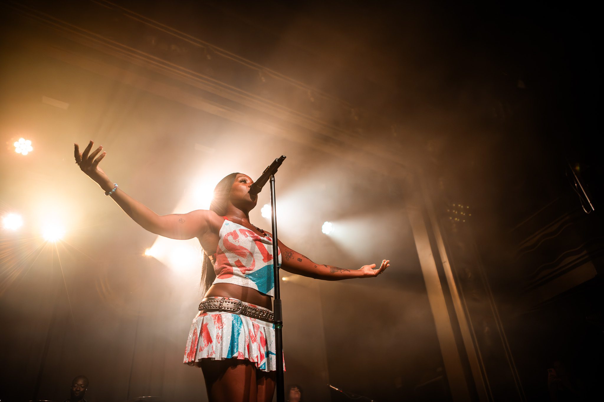A singer performing on stage with arms outstretched, illuminated by stage lights and surrounded by a smoky atmosphere.