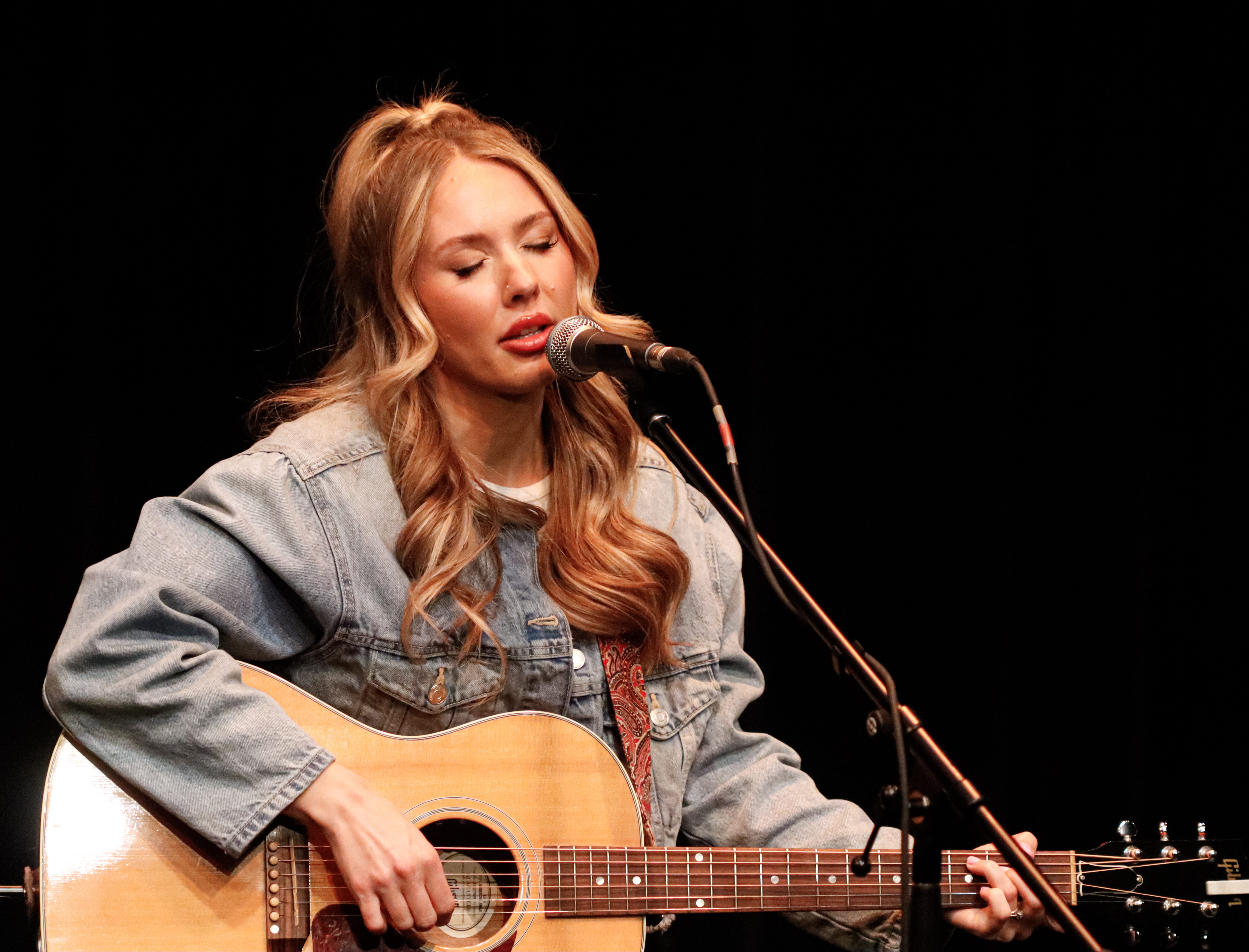 A female musician passionately singing into a microphone while playing an acoustic guitar on stage, wearing a denim jacket and appearing focused.