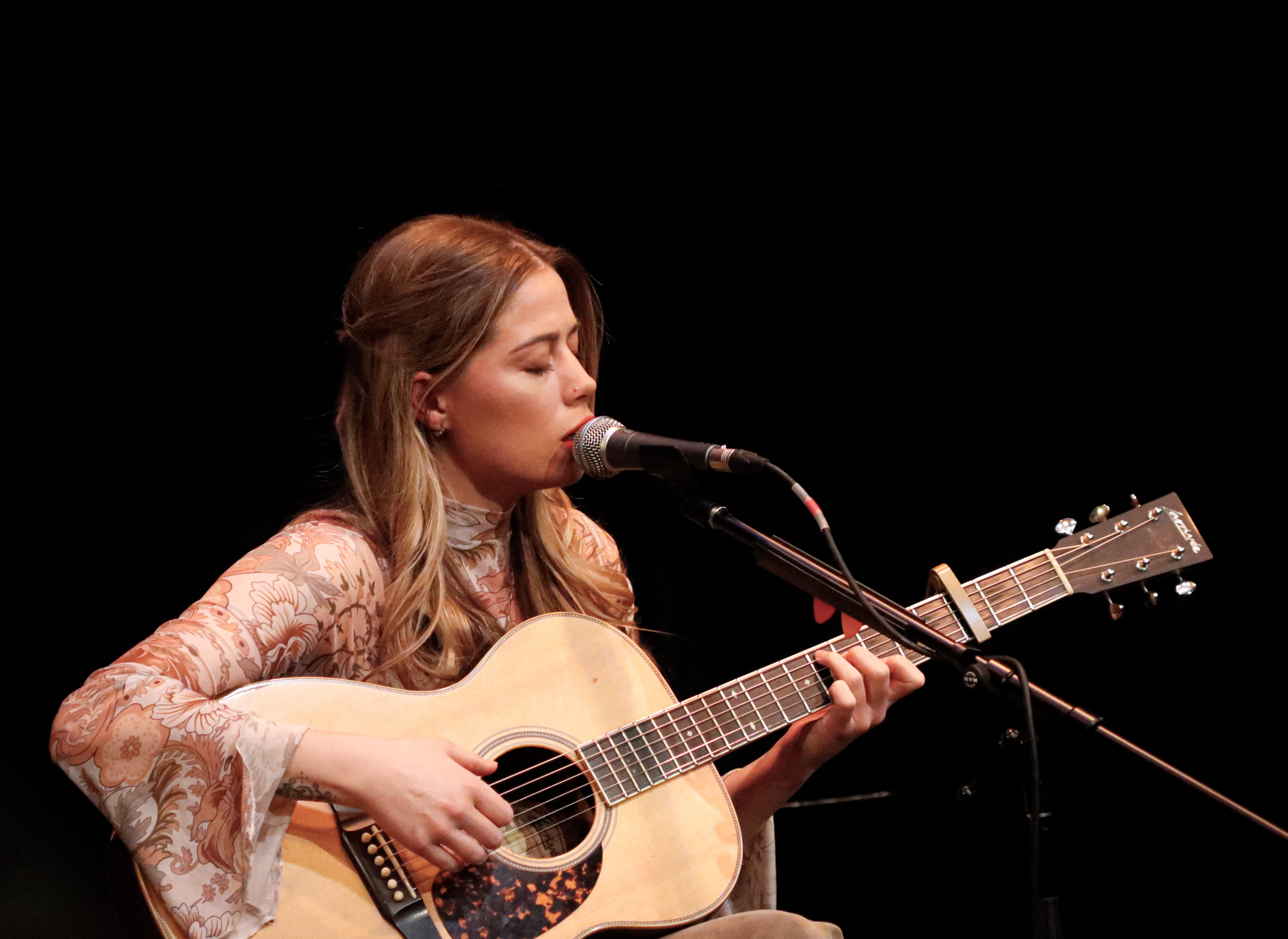 A singer-songwriter performs on stage with a guitar, focusing intently as she sings into a microphone, with a dark background highlighting her presence.
