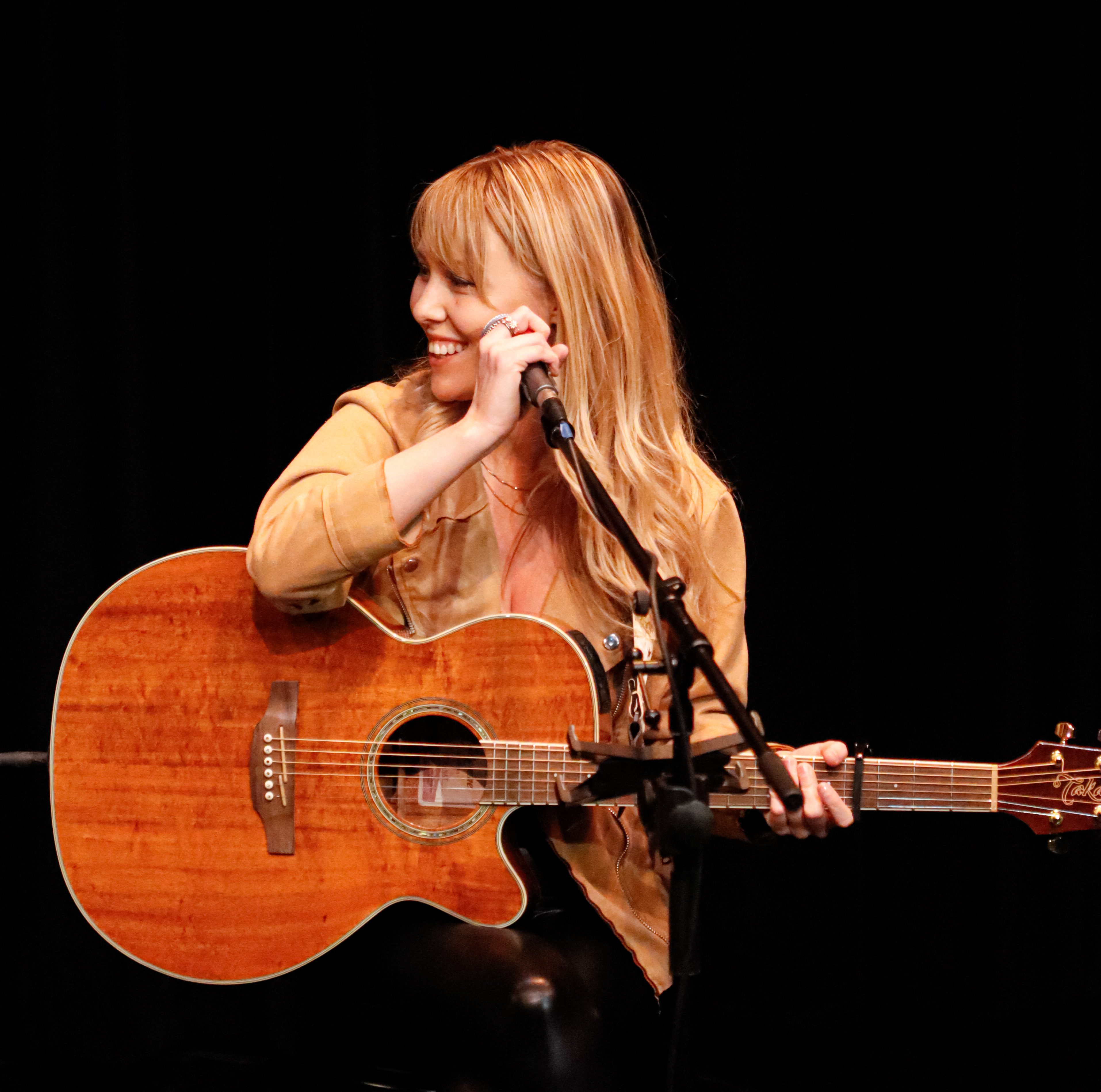 Female musician with long blonde hair seated on stage, holding a microphone and acoustic guitar, smiling while engaging with the audience.