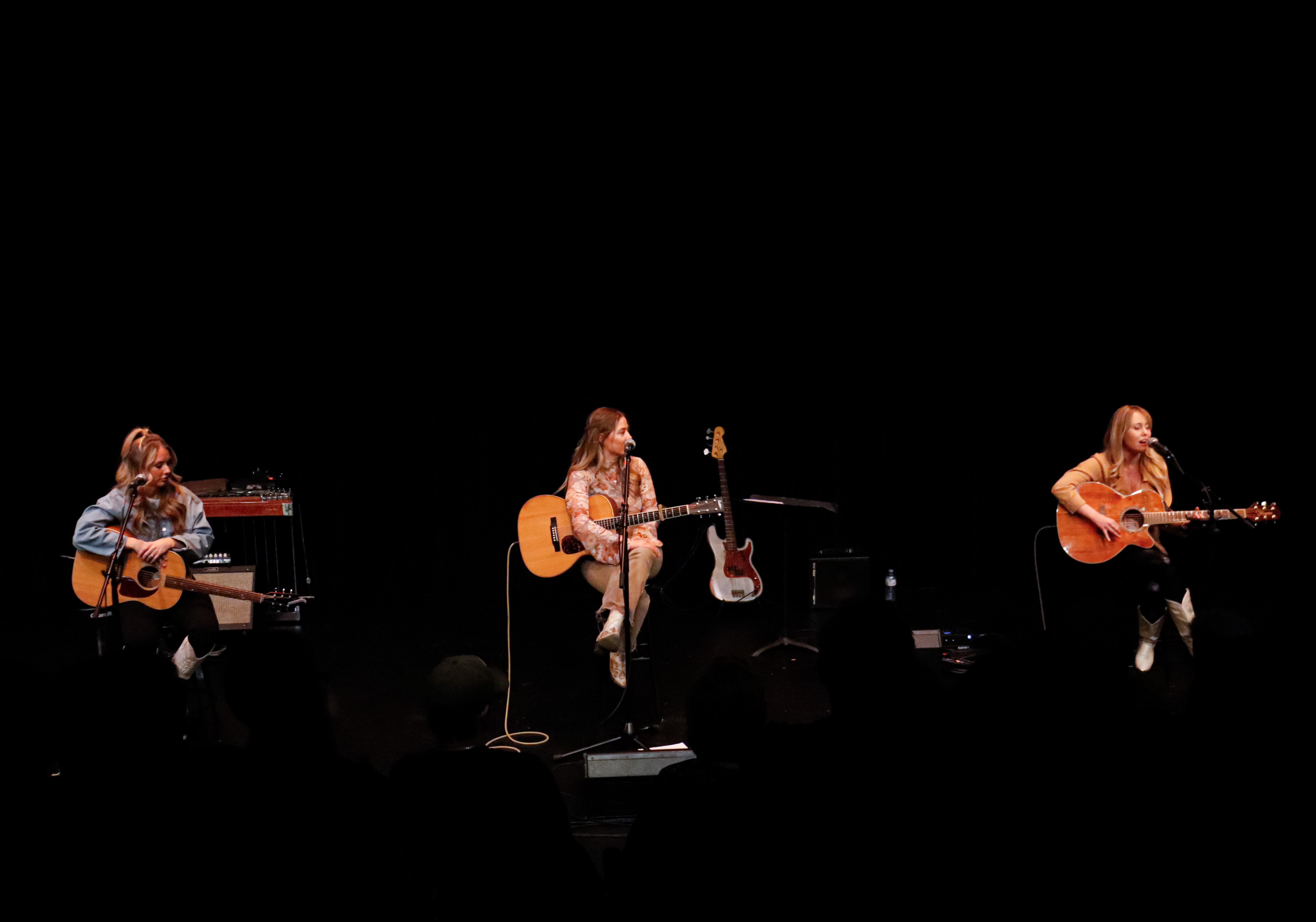 Three female artists performing on stage with acoustic guitars in a dimly lit venue.