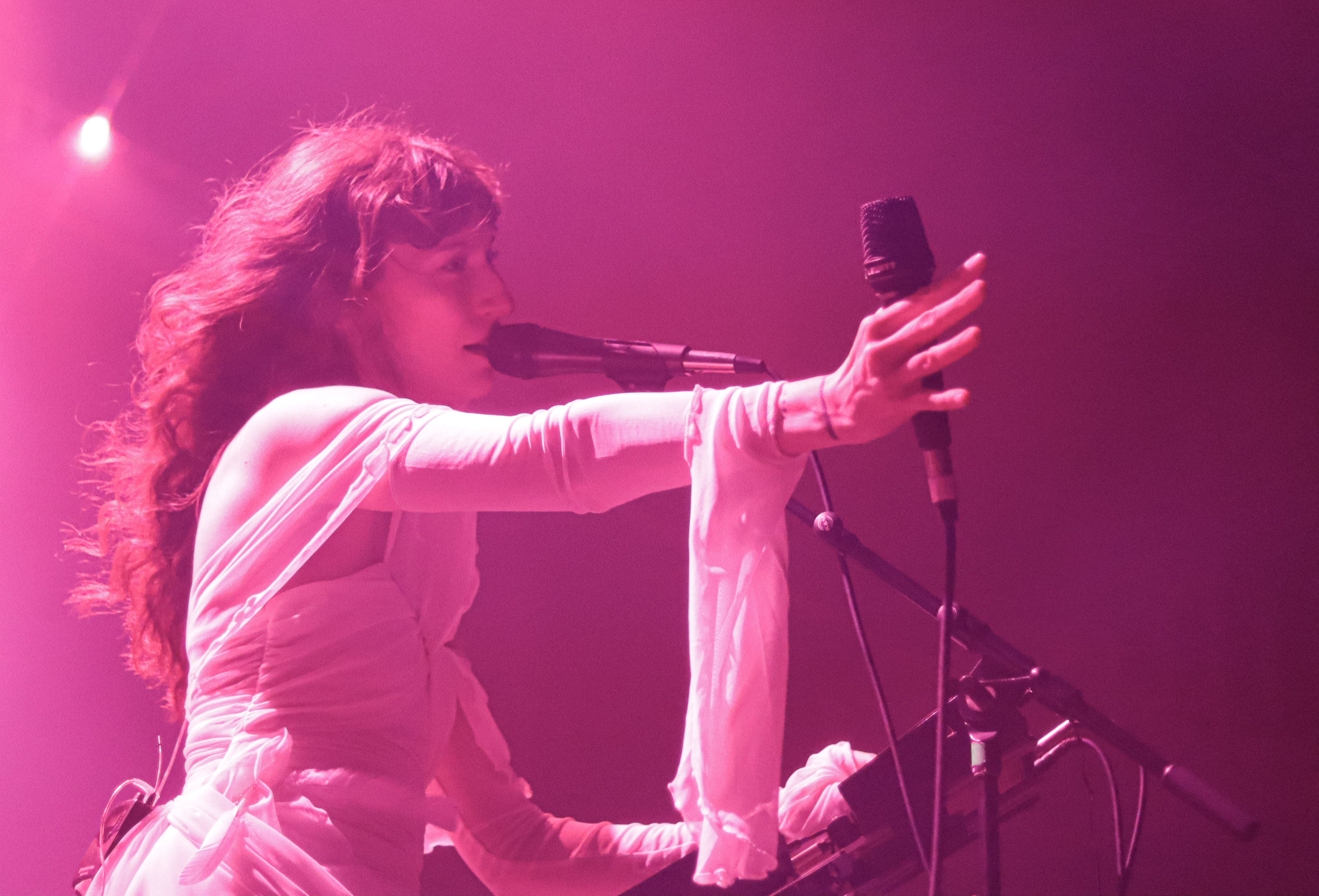 A singer performing on stage in a pink-lit environment, passionately gesturing while holding a microphone.