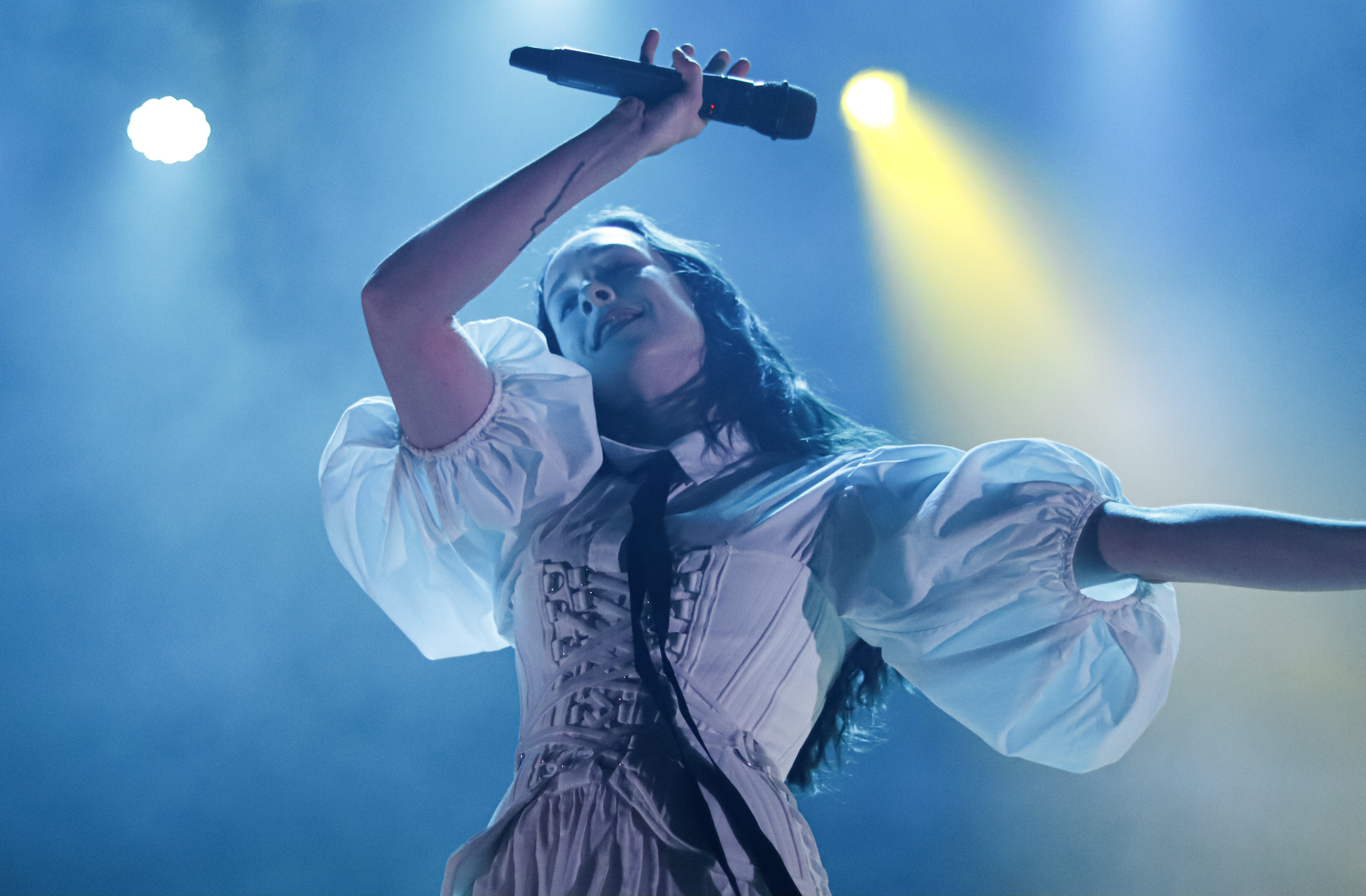 A singer performing on stage, holding a microphone, with vibrant stage lighting and smoke in the background.