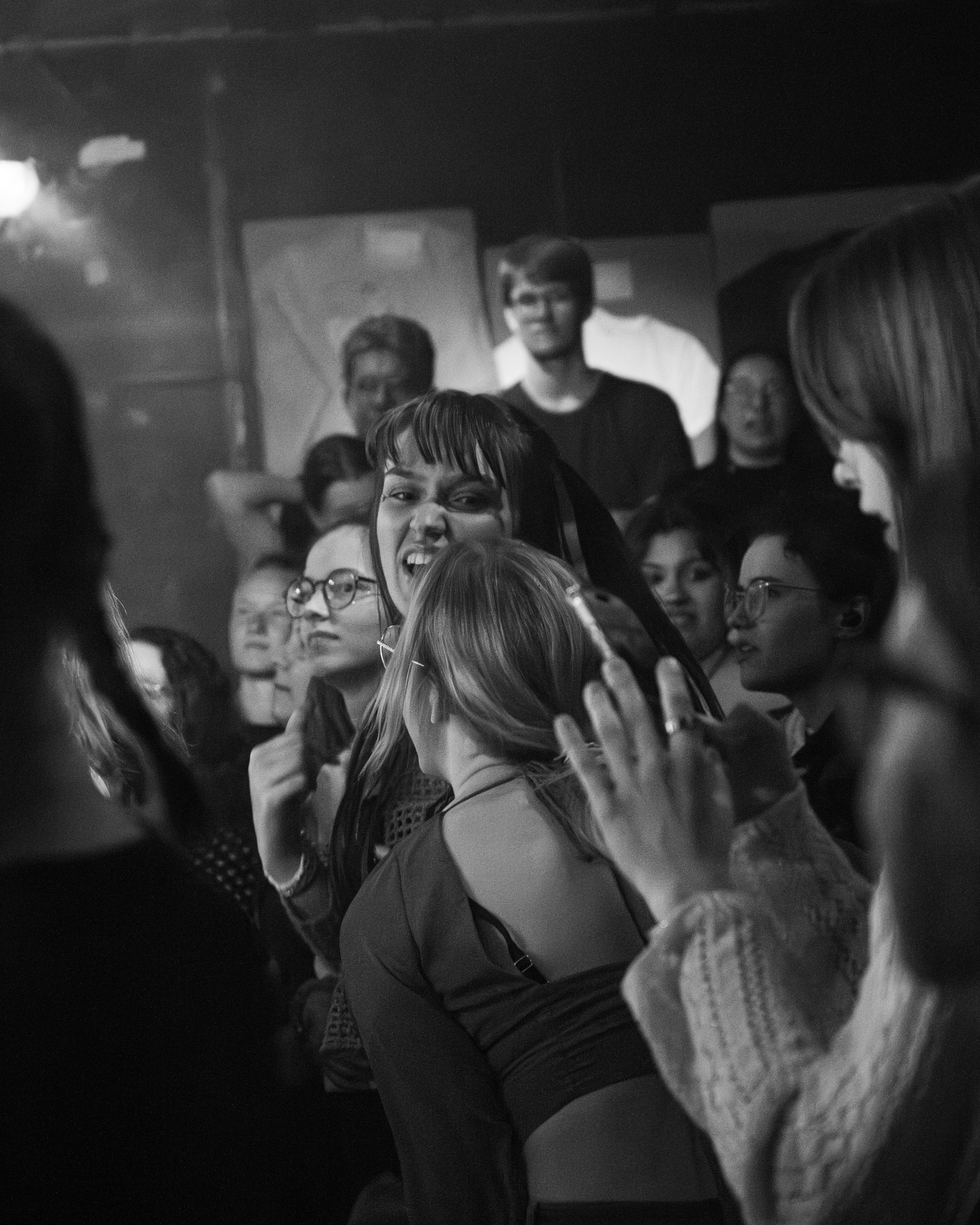 A black and white photograph of a crowd at a concert or event, featuring a diverse group of people expressing enthusiasm and engagement. Some individuals are smiling and animated, with a focus on a person in the foreground with short hair making a playful gesture.