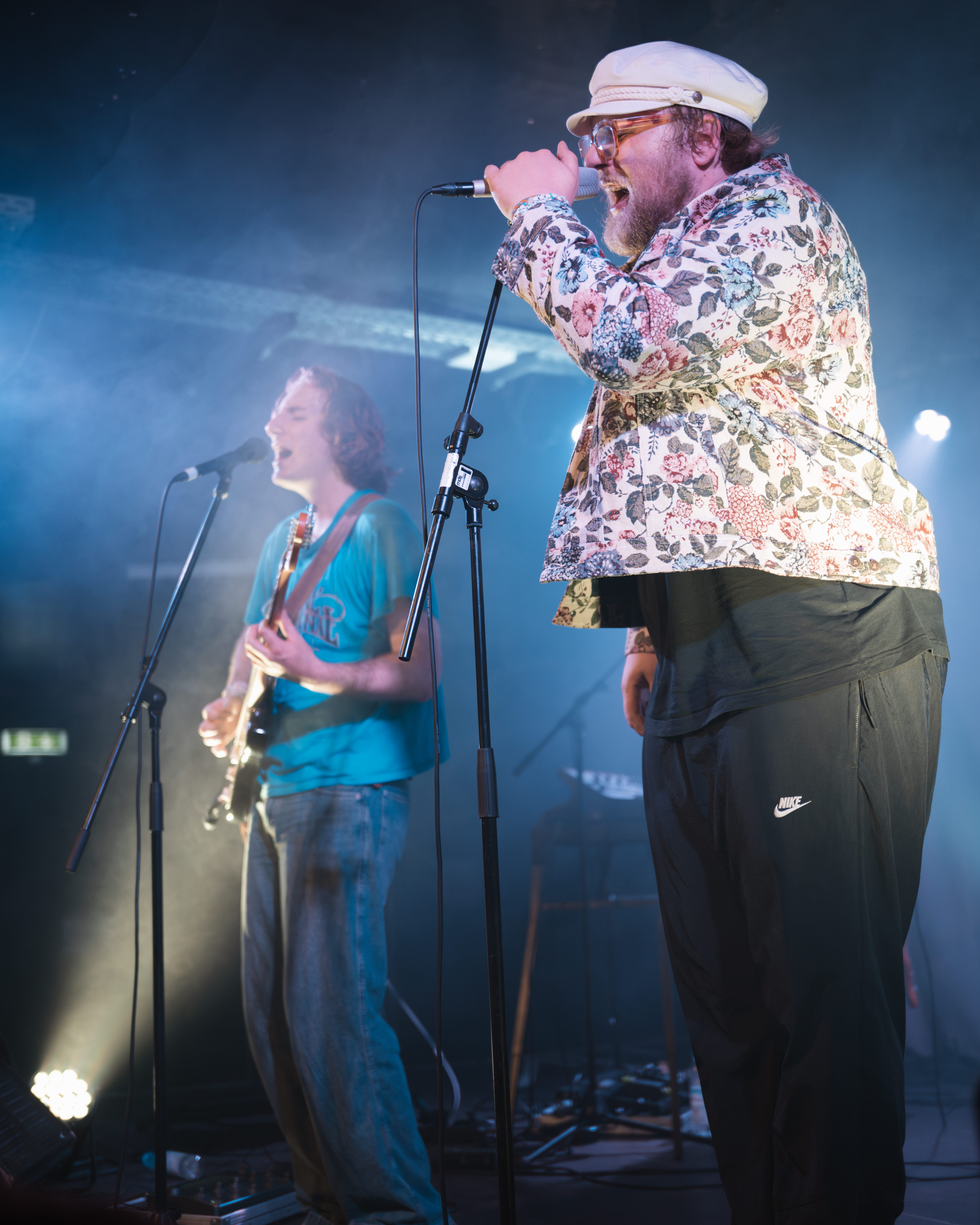 Two male musicians performing on stage. One is singing into a microphone while wearing a floral-patterned jacket and a hat, and the other is playing an electric guitar, dressed in a blue t-shirt and jeans. The stage is lit with soft lighting and a smoky atmosphere.