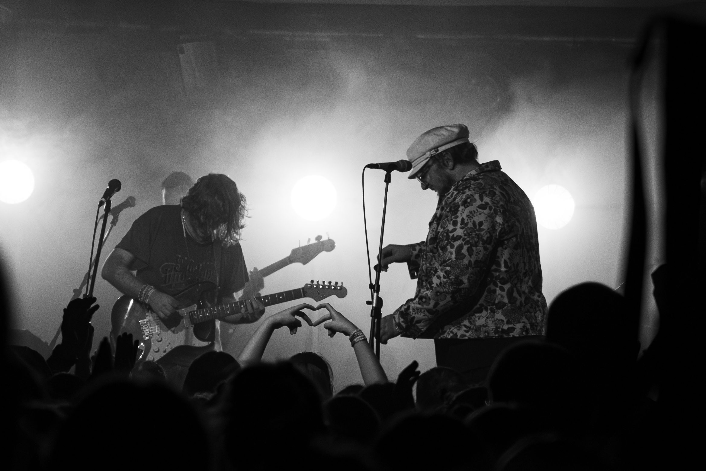 A live concert scene in black and white, featuring a guitarist playing an electric guitar and a vocalist singing into a microphone, with bright stage lights shining in the background and an engaged audience visible in the foreground.
