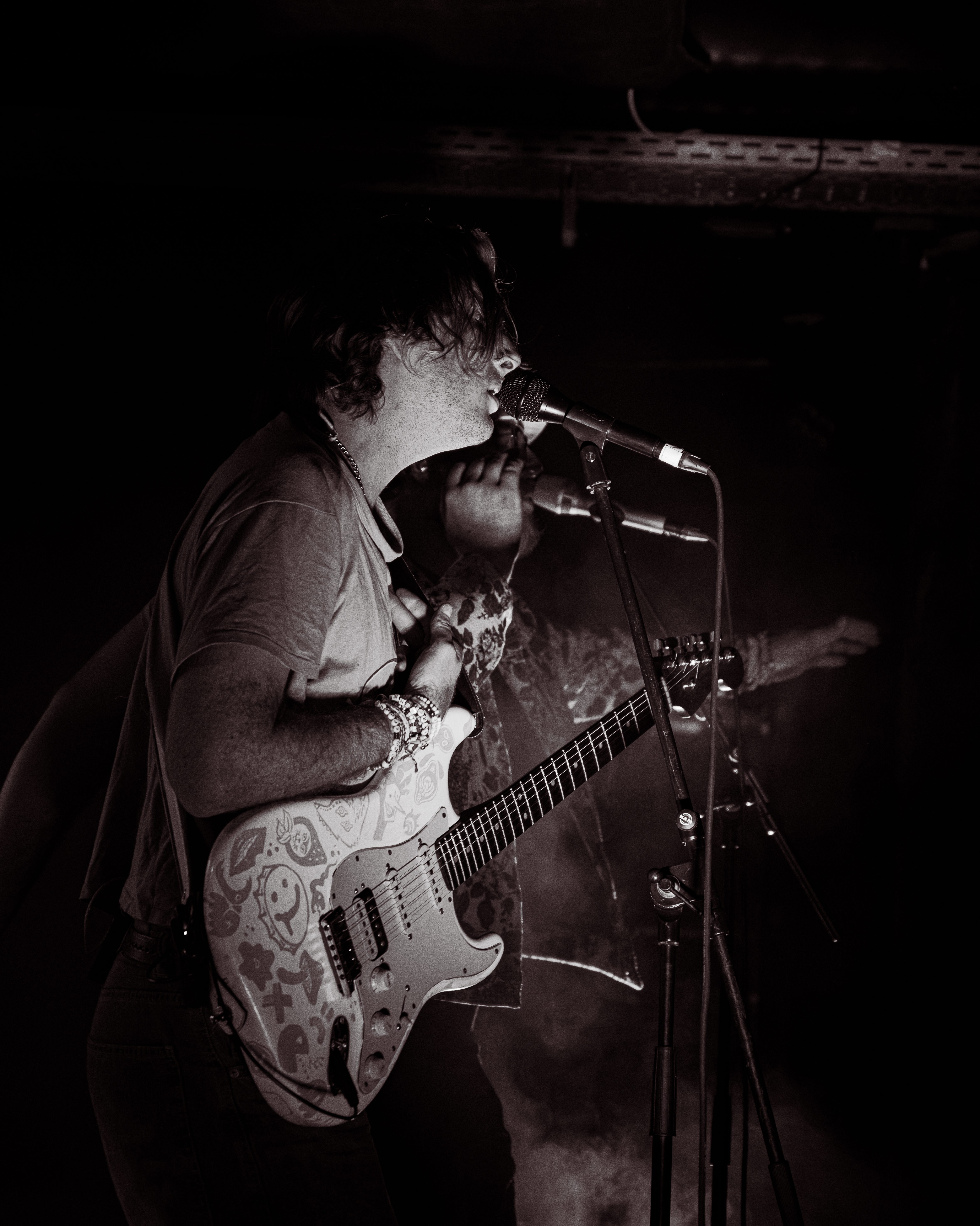 Musician performing on stage, singing into a microphone while playing a decorated electric guitar, with smoke and dim lighting in the background.