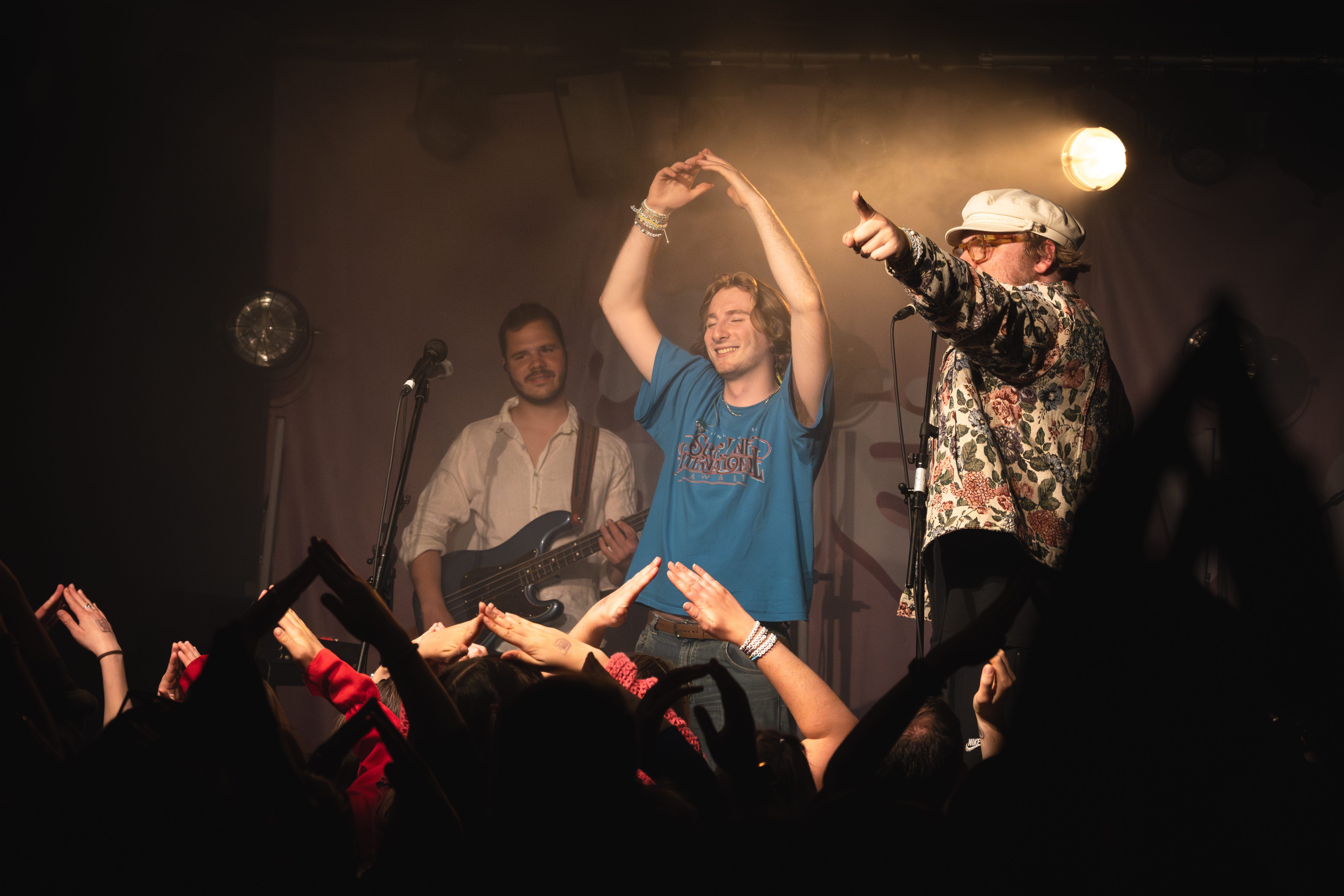 A lively concert scene with two performers on stage. One musician plays the guitar, while another, wearing a floral shirt, interacts with the audience. Fans are raising their hands in excitement, creating an energetic atmosphere.