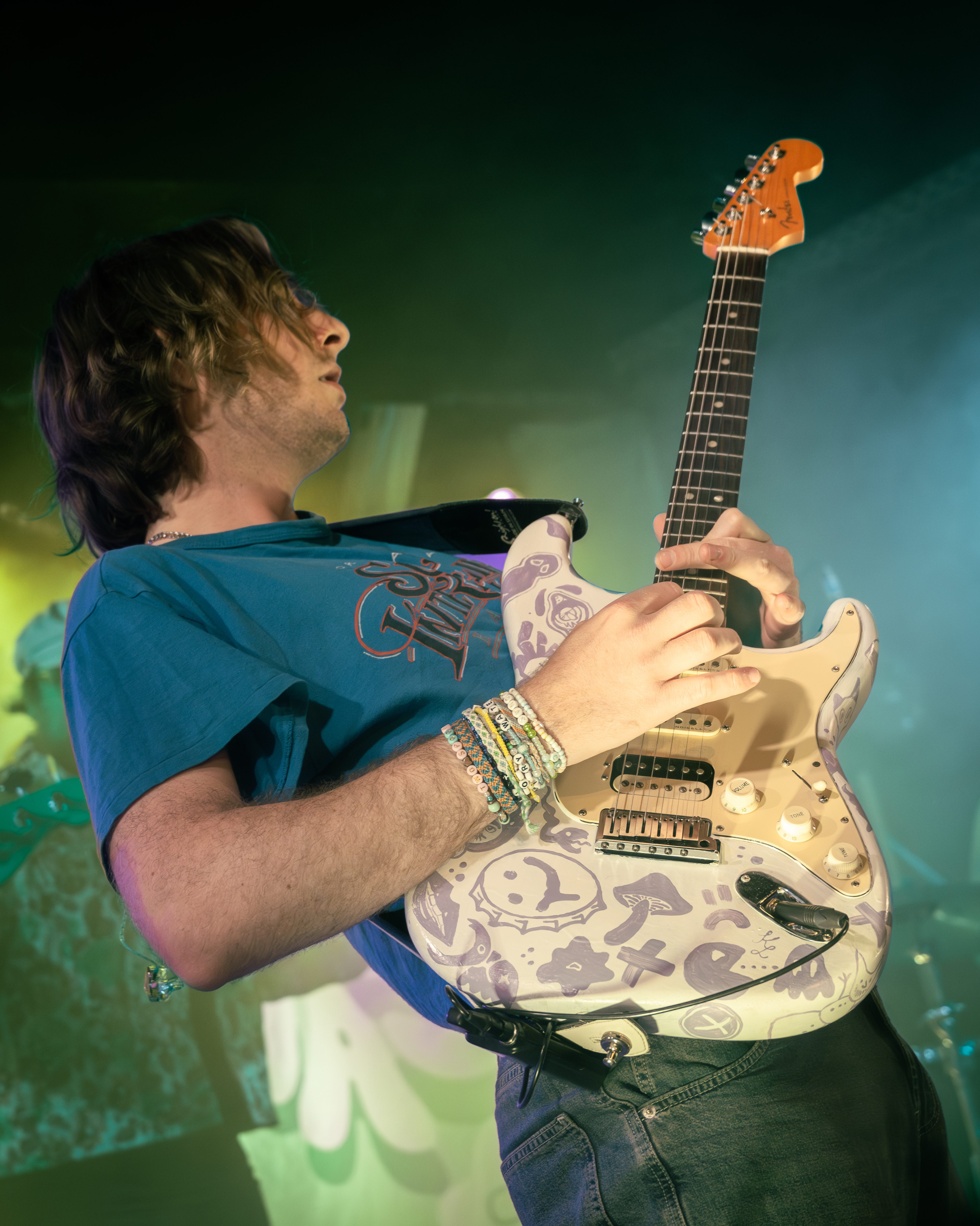 A musician performs on stage, playing a decorated electric guitar while wearing a blue t-shirt and colorful bracelets.