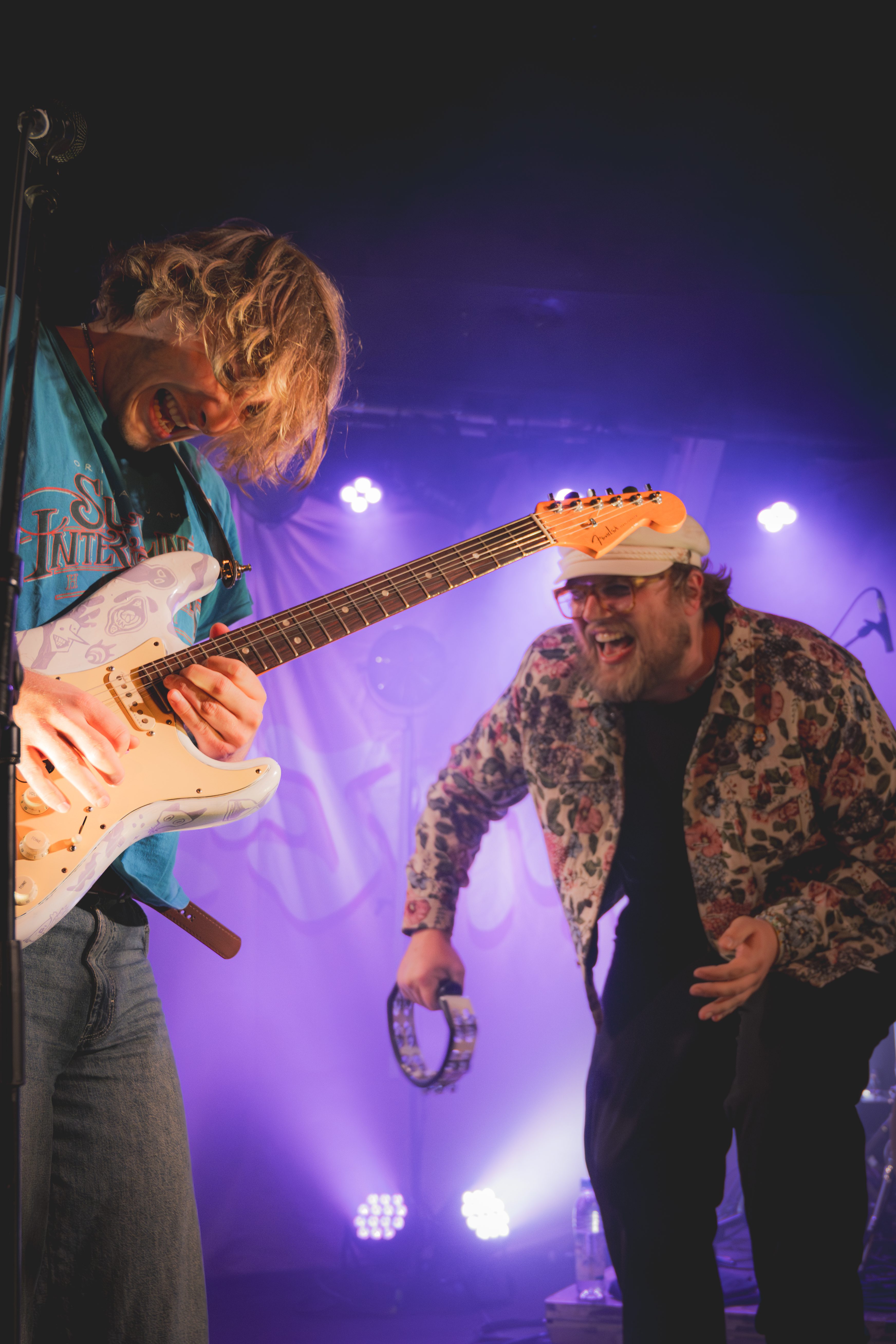 A guitarist enthusiastically plays an electric guitar while a singer joyfully holds a tambourine on stage, with colorful stage lights in the background.