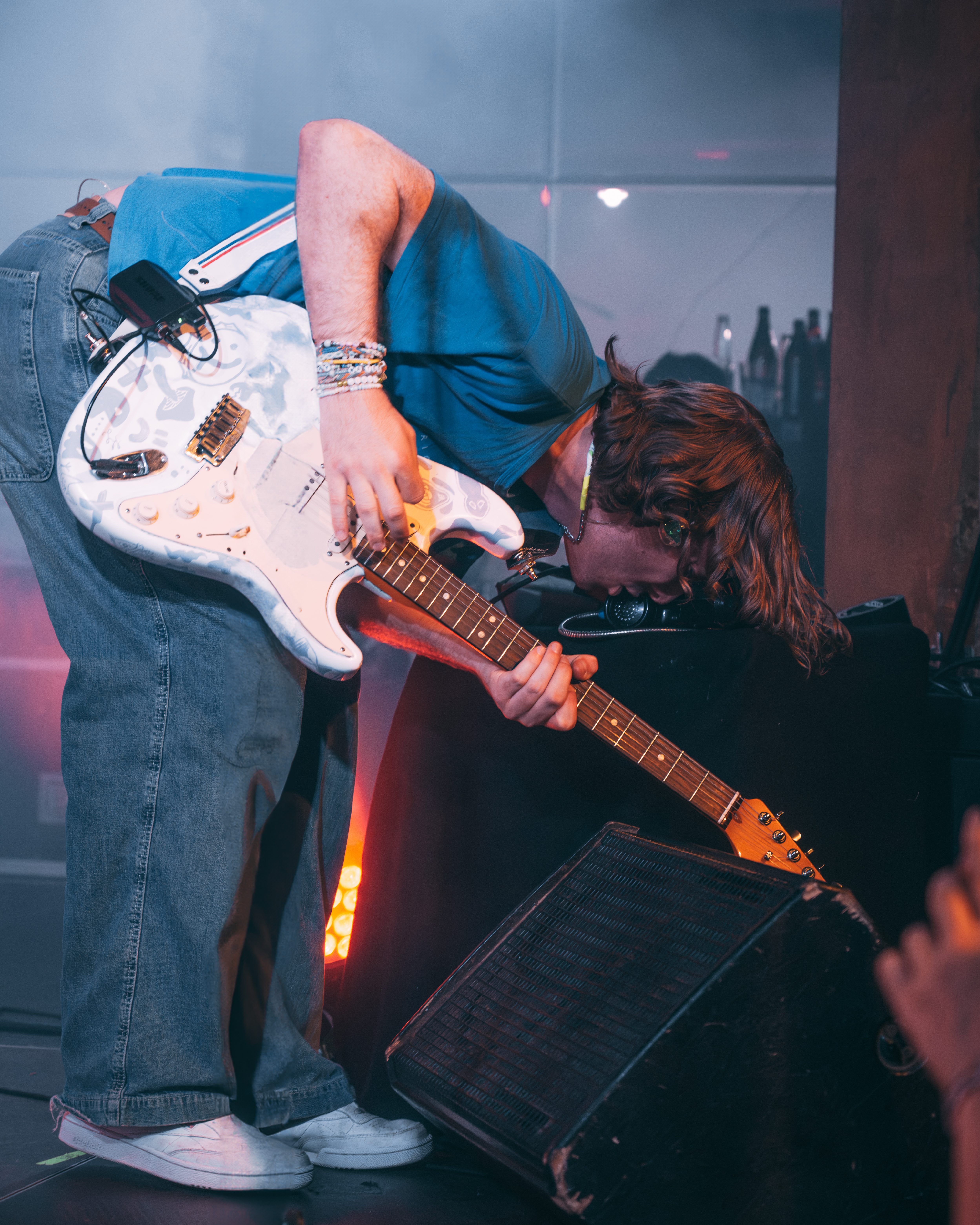 A musician bending down to play an electric guitar, surrounded by colorful stage lighting and smoke.