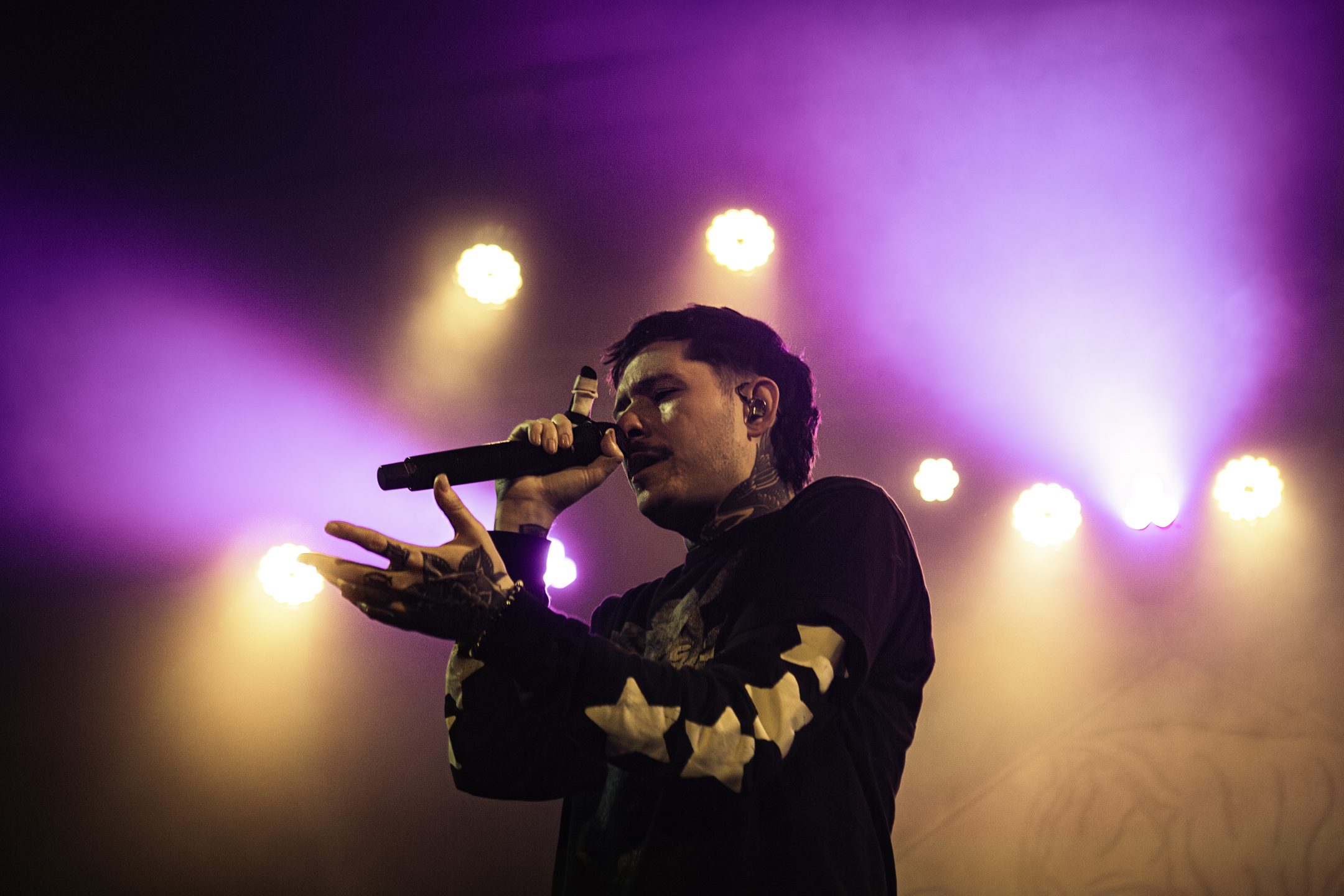 A performer singing into a microphone on stage, illuminated by purple and white stage lights, wearing a black shirt with white designs.