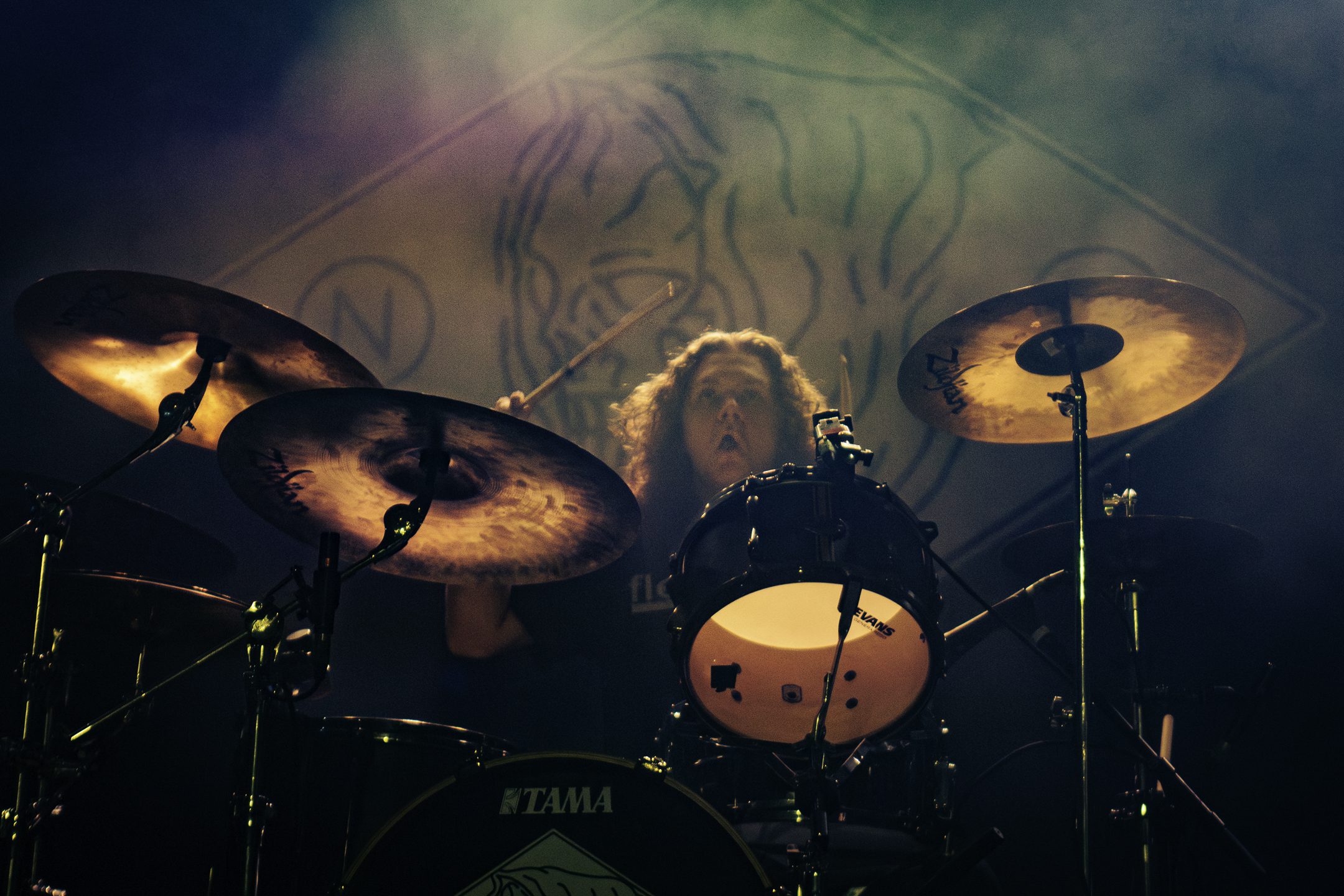 A drummer performing on stage, focused and energetic, with drumsticks raised and a dramatic smoky backdrop.