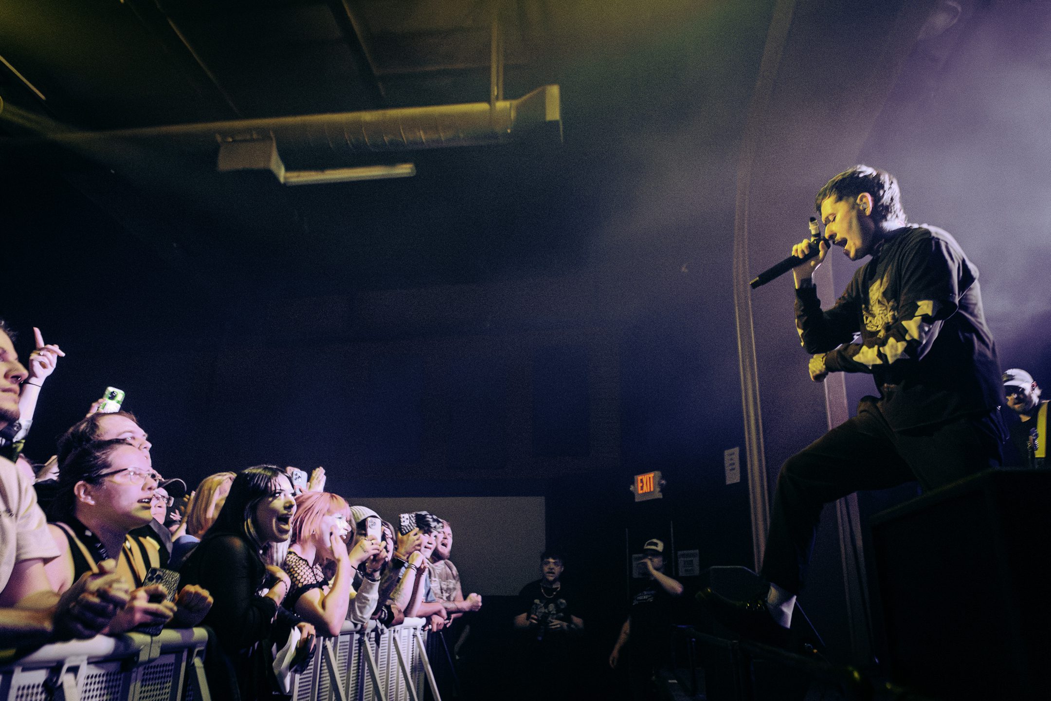A musical performer singing into a microphone on stage, with a captivated audience in the foreground, some fans visibly excited and holding their phones to capture the moment.
