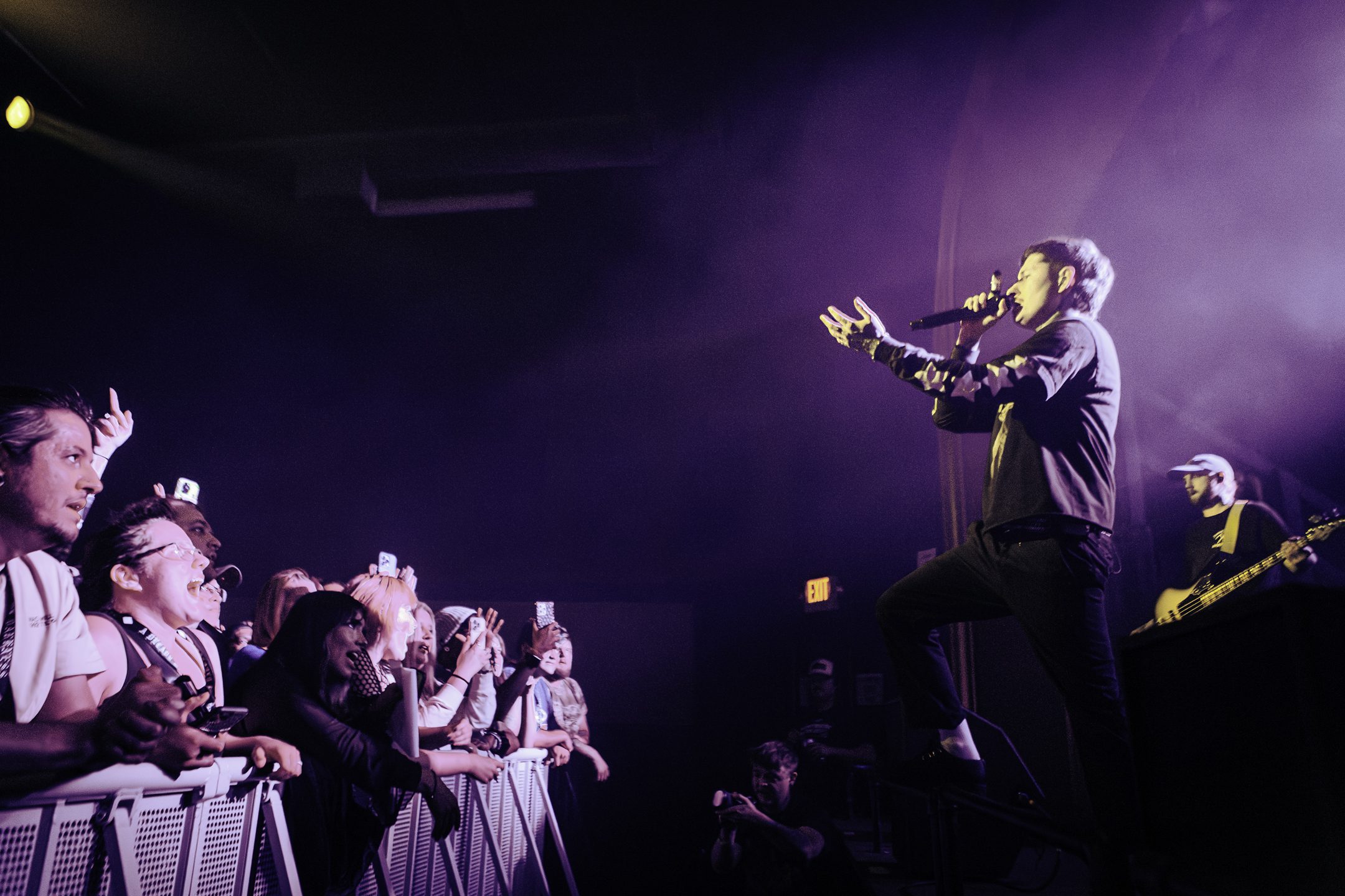 A live performance scene featuring a male singer passionately engaging with the audience at a concert, with fans cheering and recording on their phones in a vibrant atmosphere.