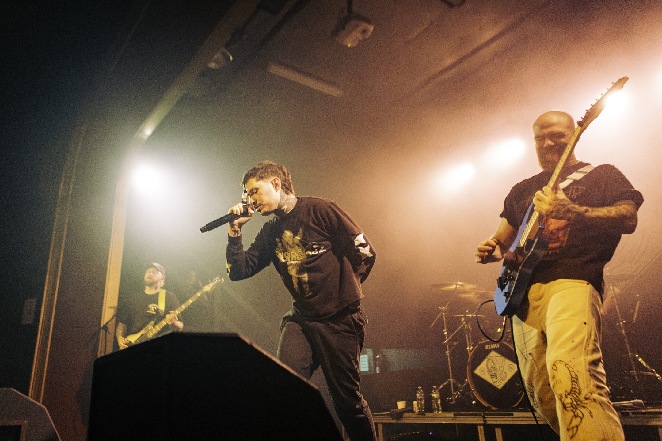 Musician performing on stage with microphone, accompanied by a guitarist and other band members in the background under dramatic lighting.